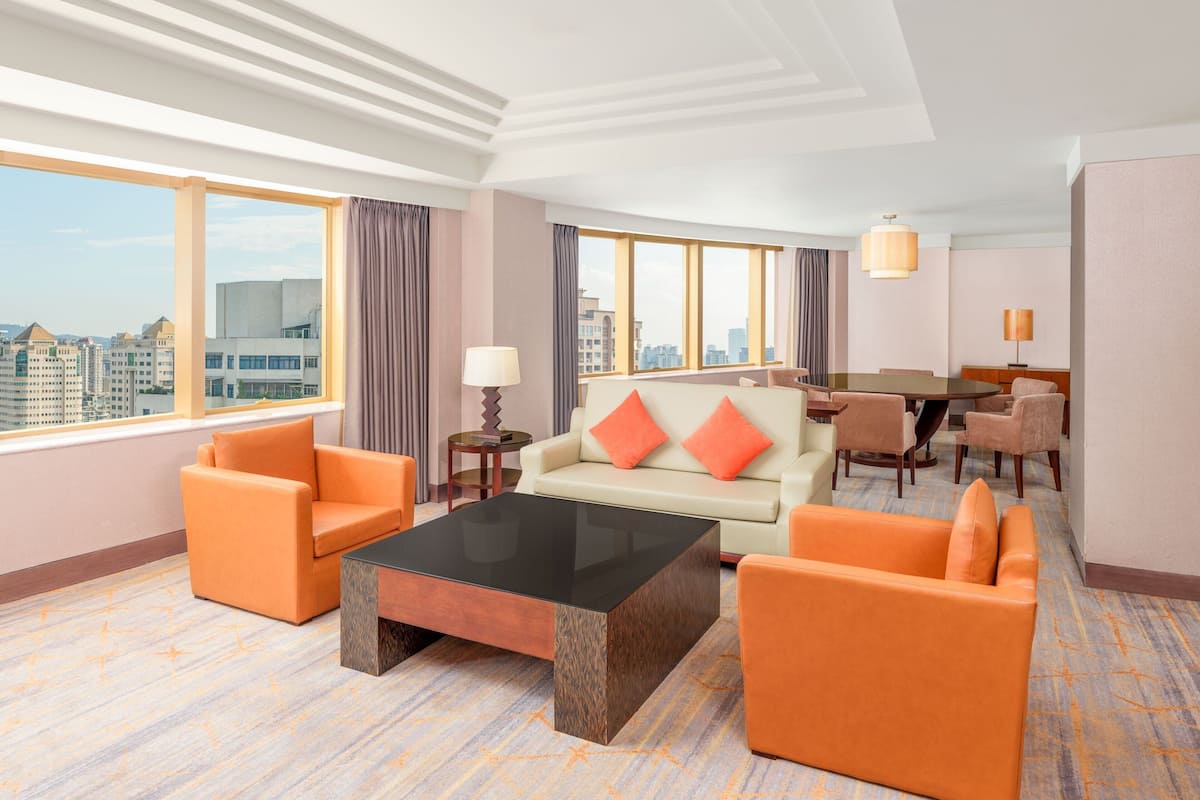 A hotel room with a large window, a sofa, two armchairs, a coffee table, and a dining area with chairs.