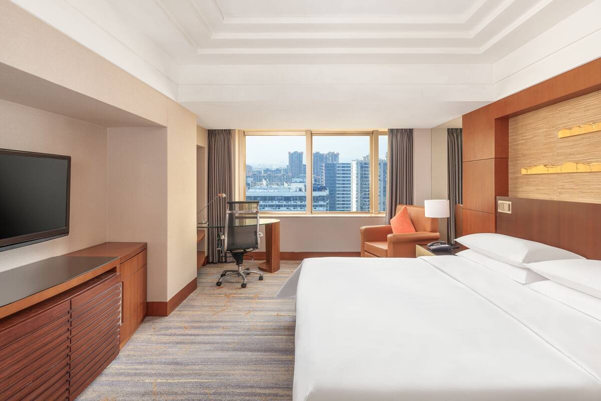 A hotel room with a large bed, a desk, a chair, a TV, and a window with a city view.