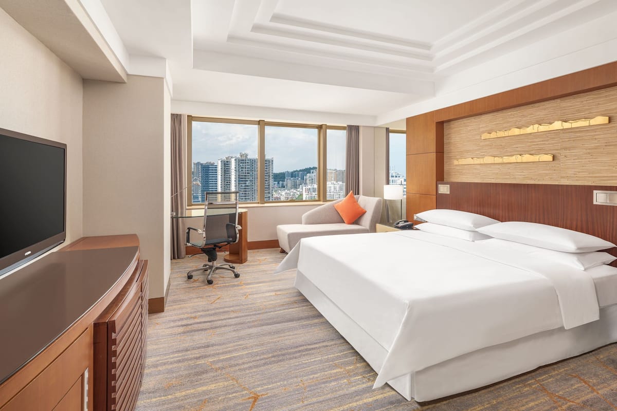 A hotel room with a large bed, a desk with a chair, a television, and a city view through large windows.
