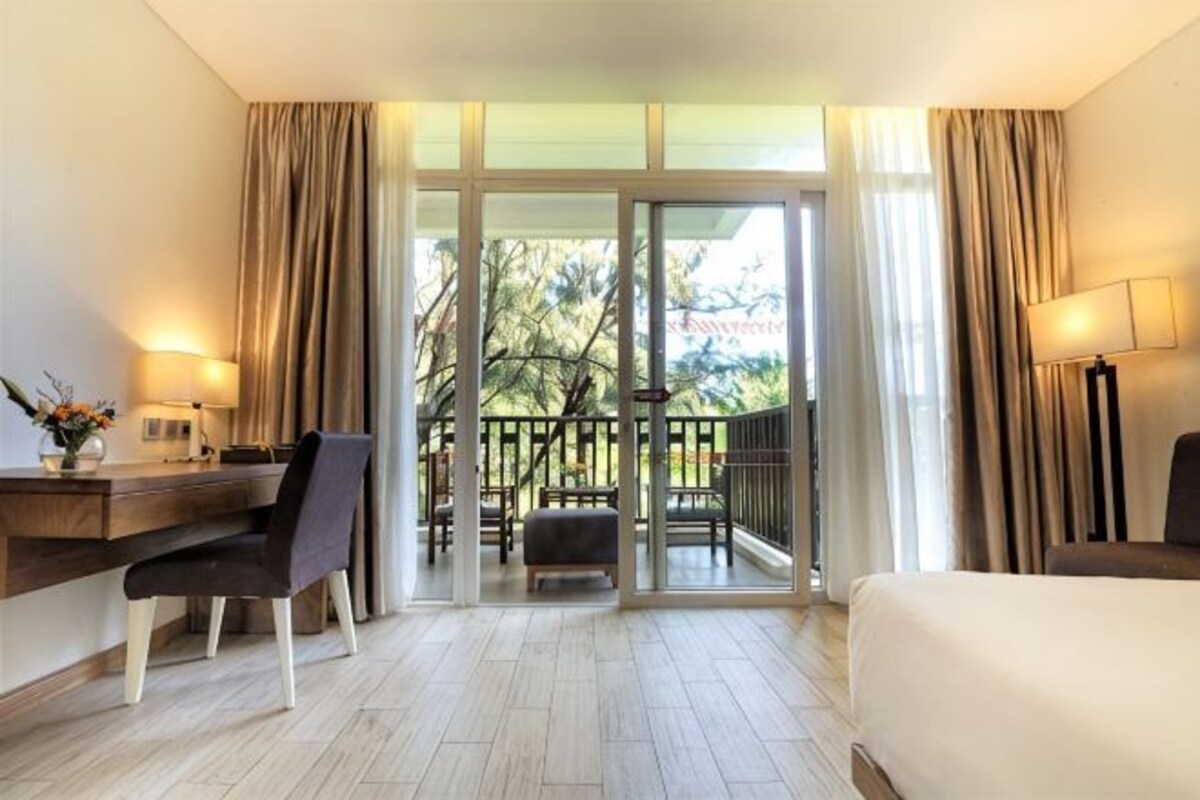 A hotel room with a bed, desk, chair, and a balcony with a view of trees.