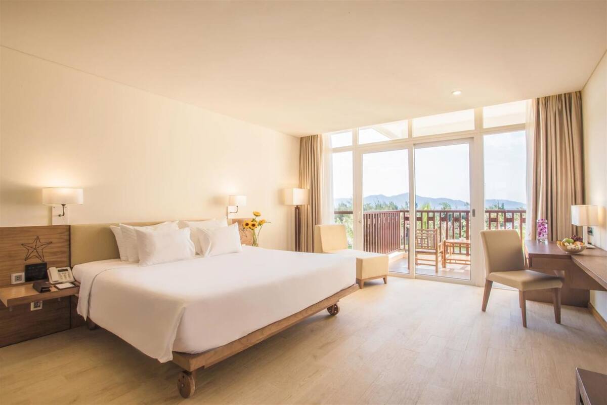 A hotel room with a large bed, a desk, a chair, and a balcony with a view.