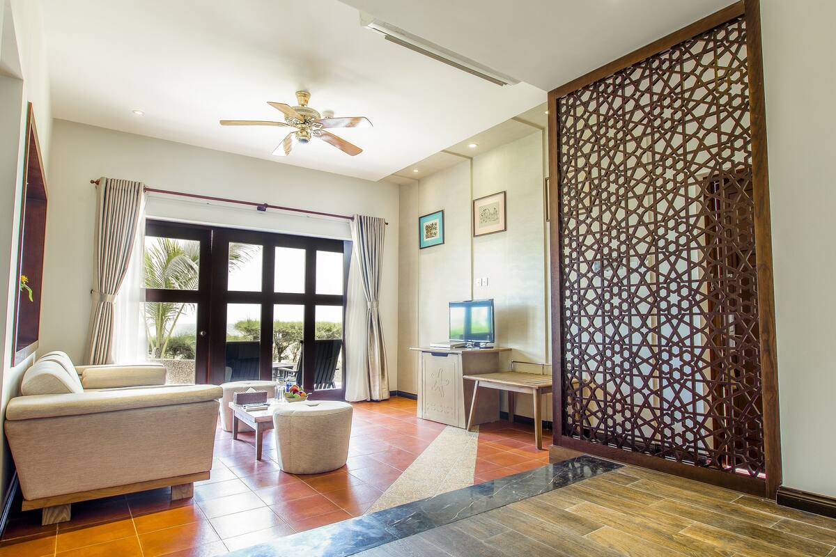 A spacious living area with a sofa, a coffee table, and a TV. There is a decorative wooden screen and a ceiling fan.