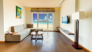 A modern hotel room with a sofa, a small round table with a vase of flowers, a flat-screen TV mounted on the wall, and a large window with a beach view.