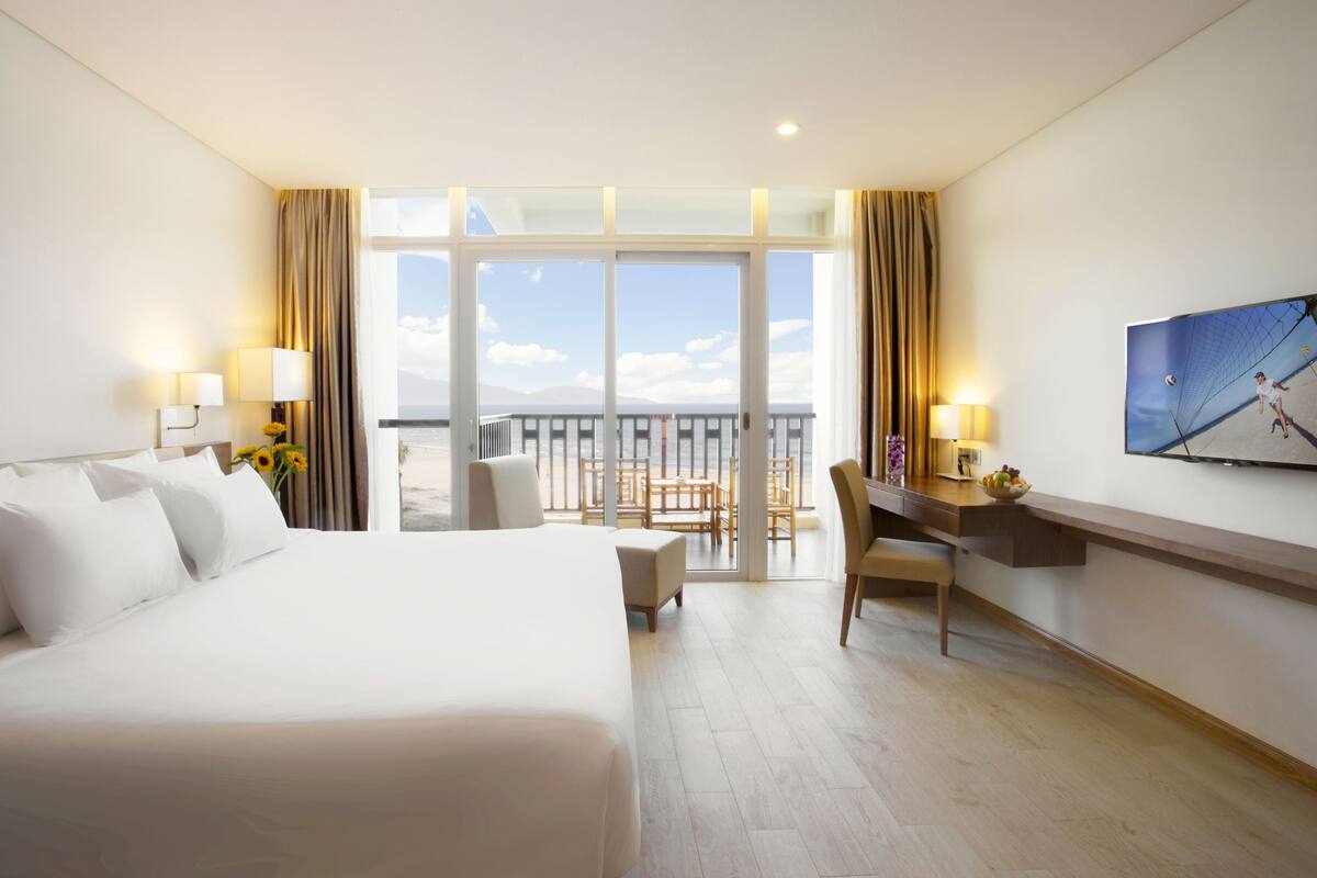 A hotel room with a large bed, a desk, a chair, a TV, and a balcony with a view of the beach.