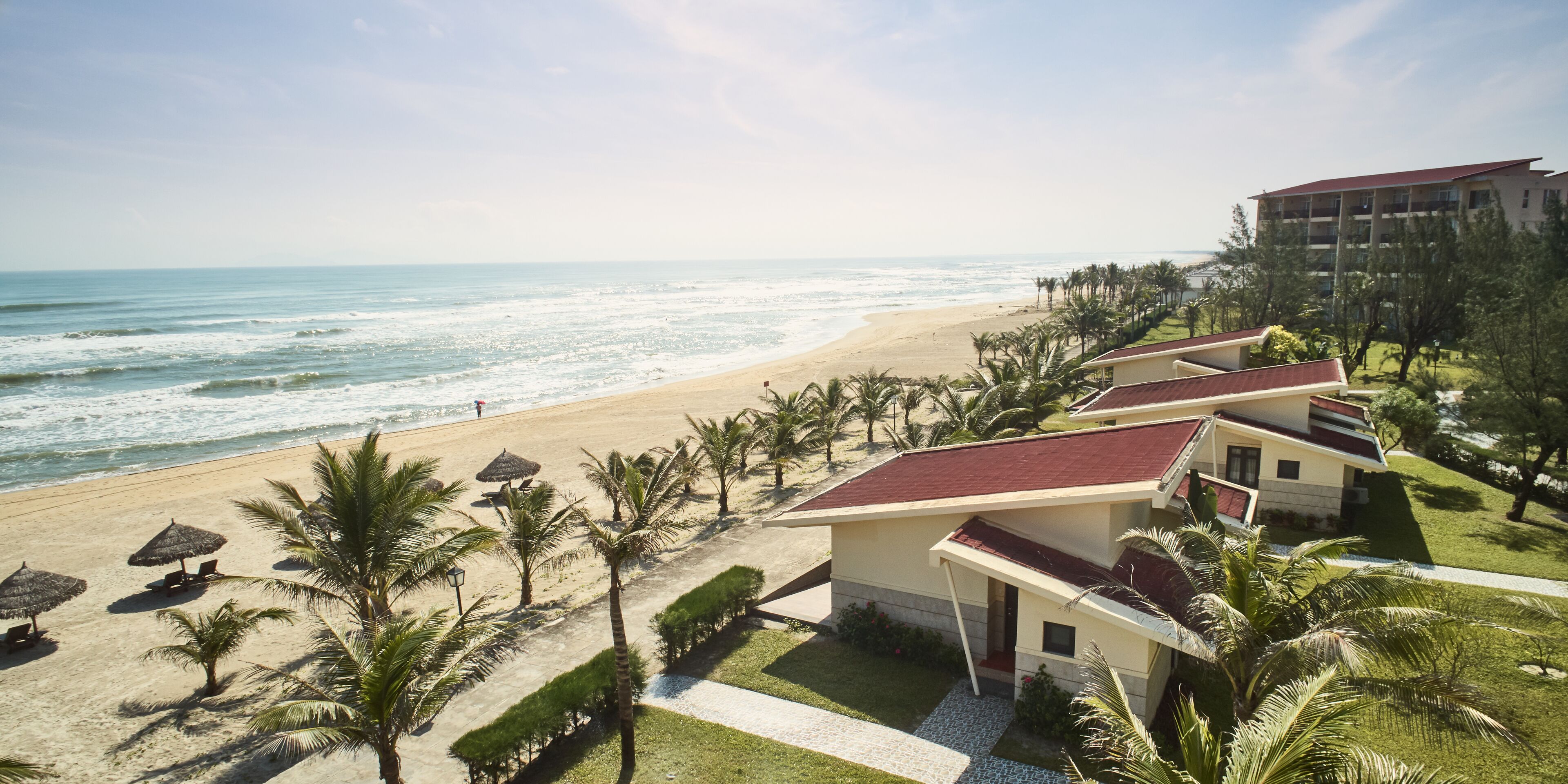 Un hotel frente a la playa con varias habitaciones, una playa de arena y palmeras.