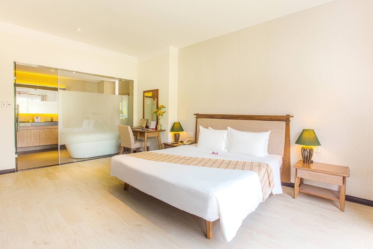 A modern hotel room with a large bed, a desk, and a bathroom visible through a glass partition.