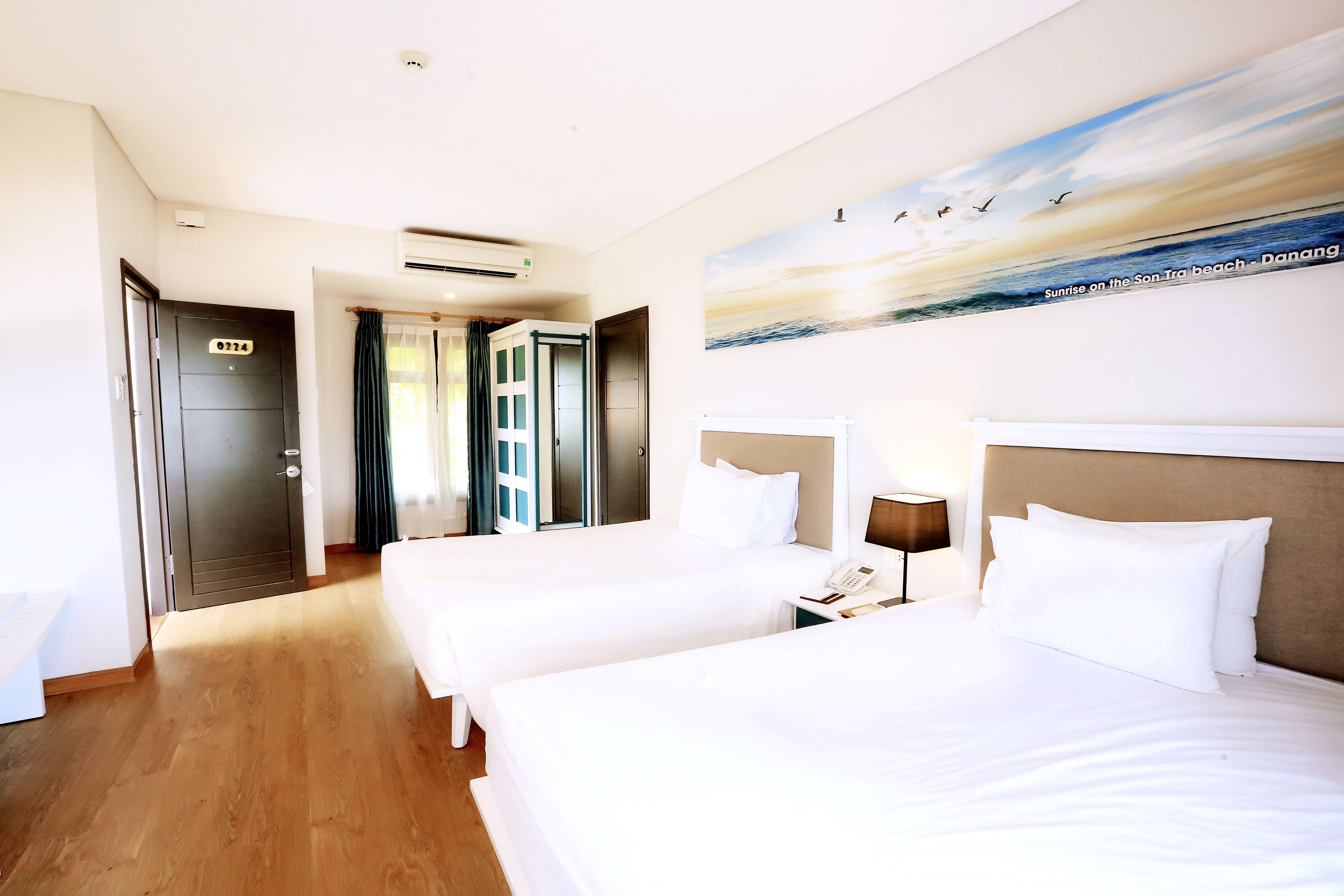 A hotel room with two beds, a painting of a beach, and a door with a number.