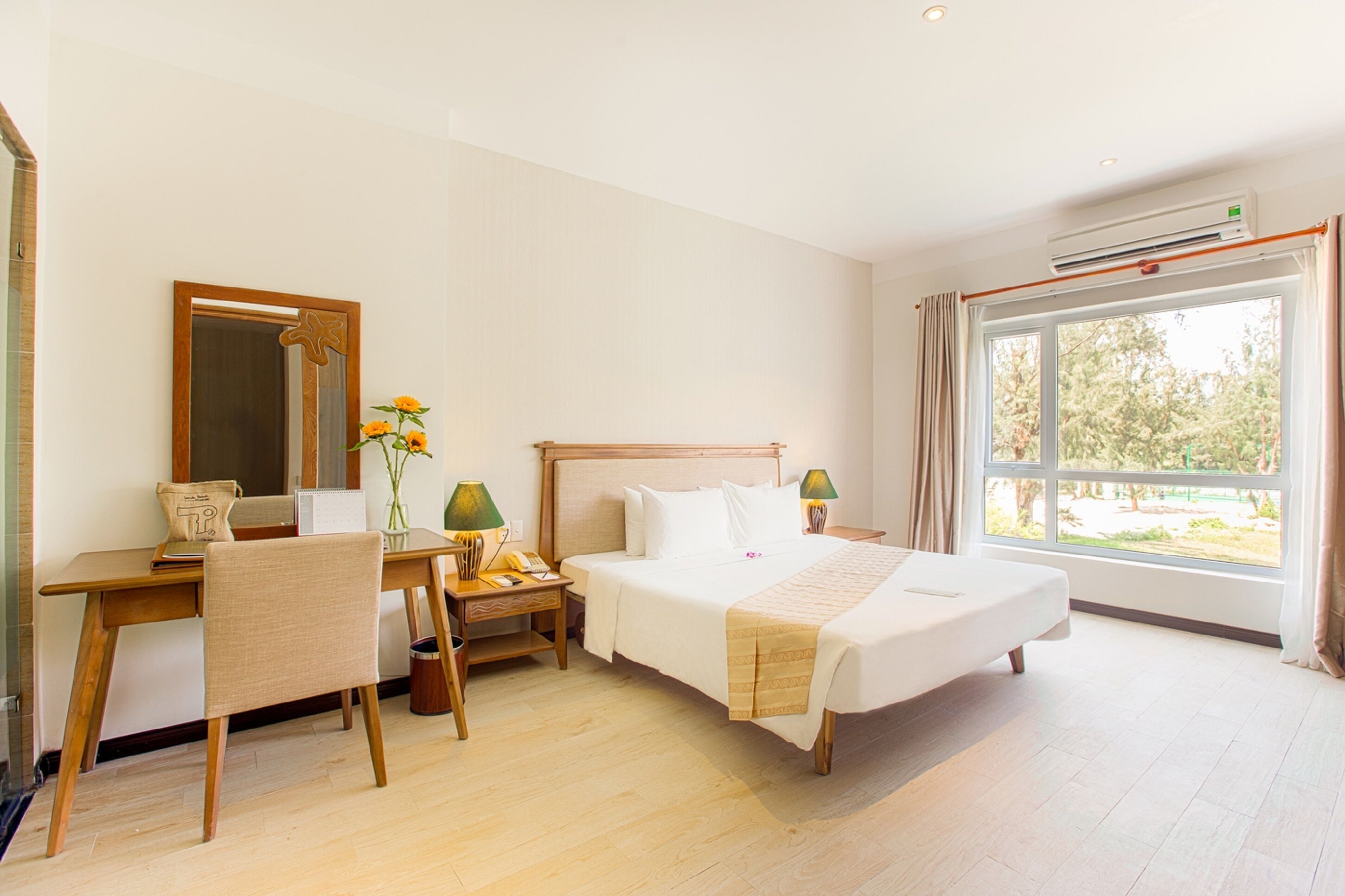A hotel room with a bed, a desk, a chair, a mirror, a bedside table with lamps, and a window with a view of trees.