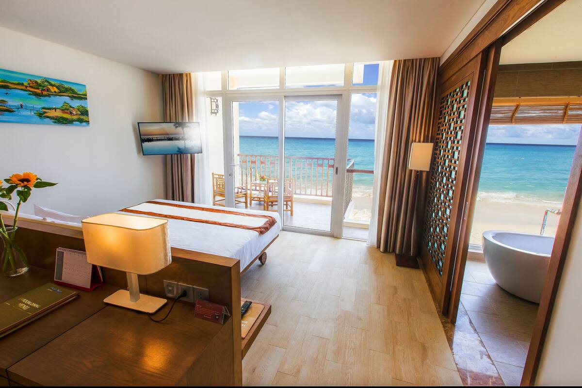 A hotel room with a bed, a desk with a lamp, a TV, a calendar, a view of the beach, and a bathroom with a large bathtub.