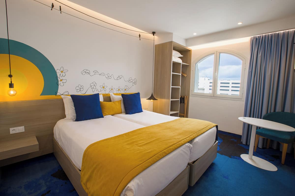 A hotel room with a bed, bedside table, lamp, chair, and a view of the city.