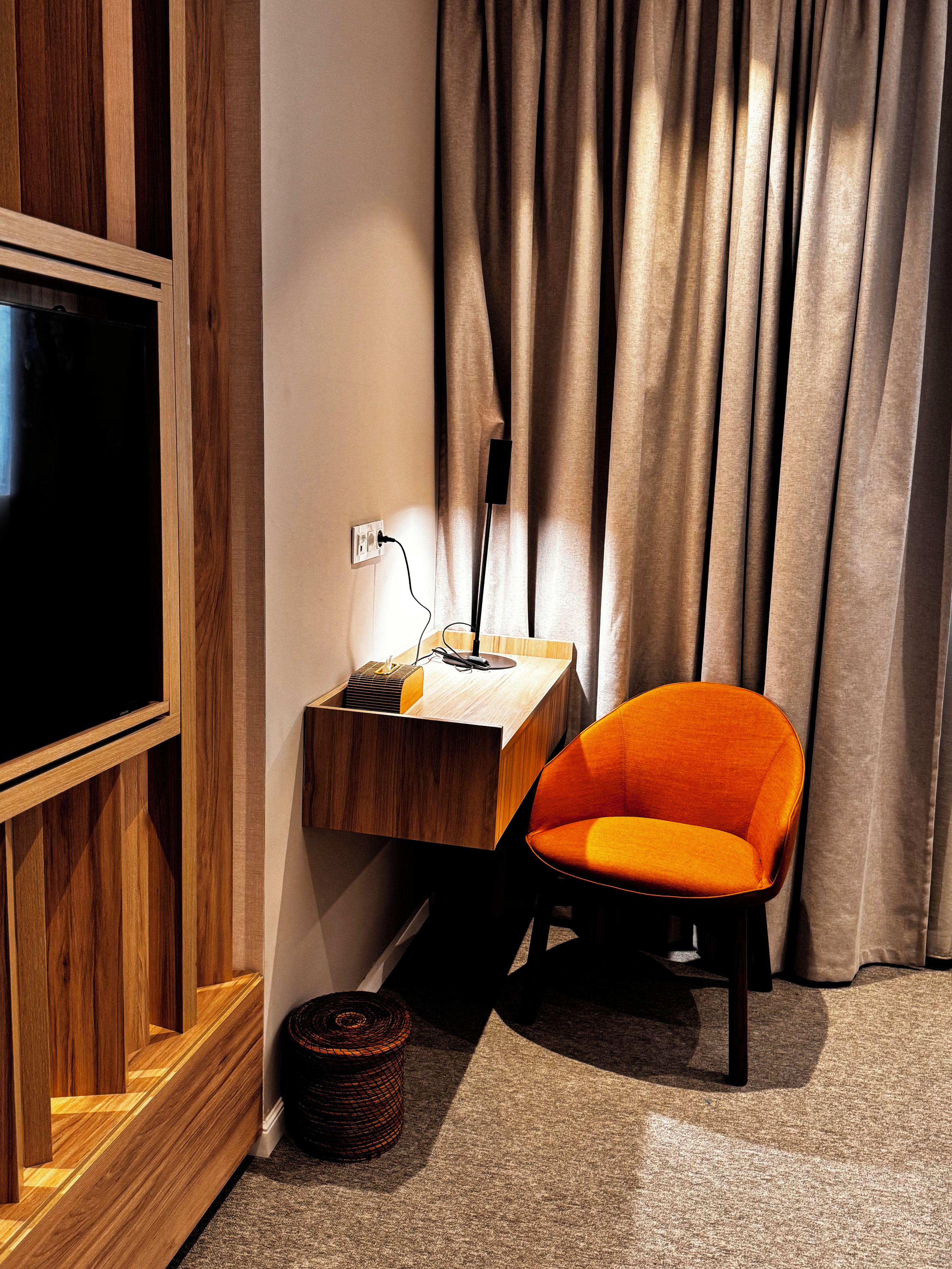 Una habitación moderna con un mueble de televisión de madera, un escritorio con lámpara, una silla naranja y un amplio ventanal con cortinas.