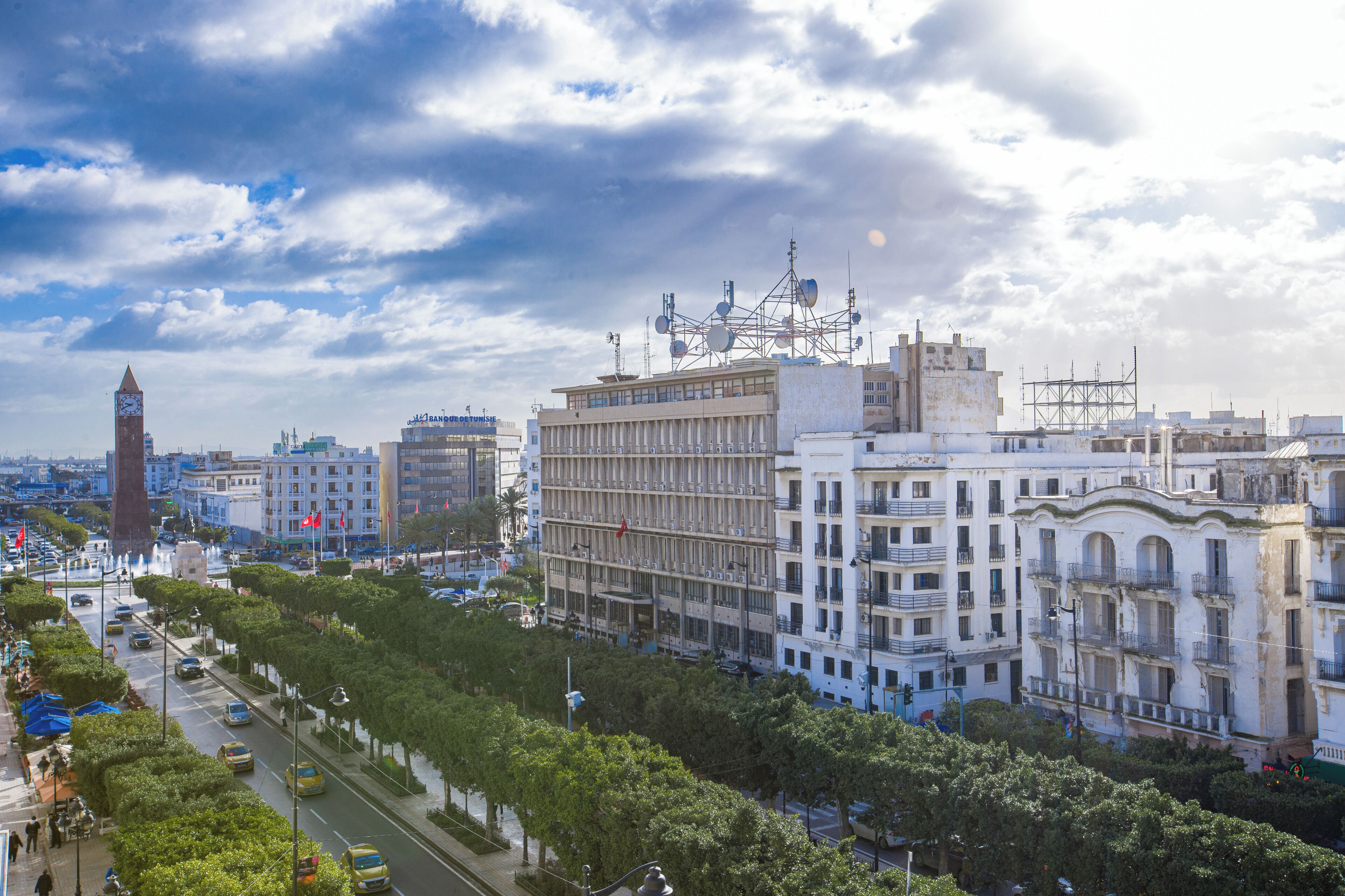 Un paisaje urbano con una torre de reloj destacada, edificios de varios pisos y una calle concurrida con vehículos y peatones.