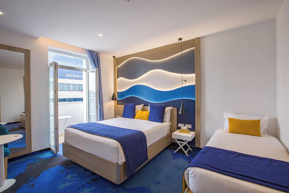 A modern hotel room with a large bed, a smaller bed, a desk, and a view of the city.