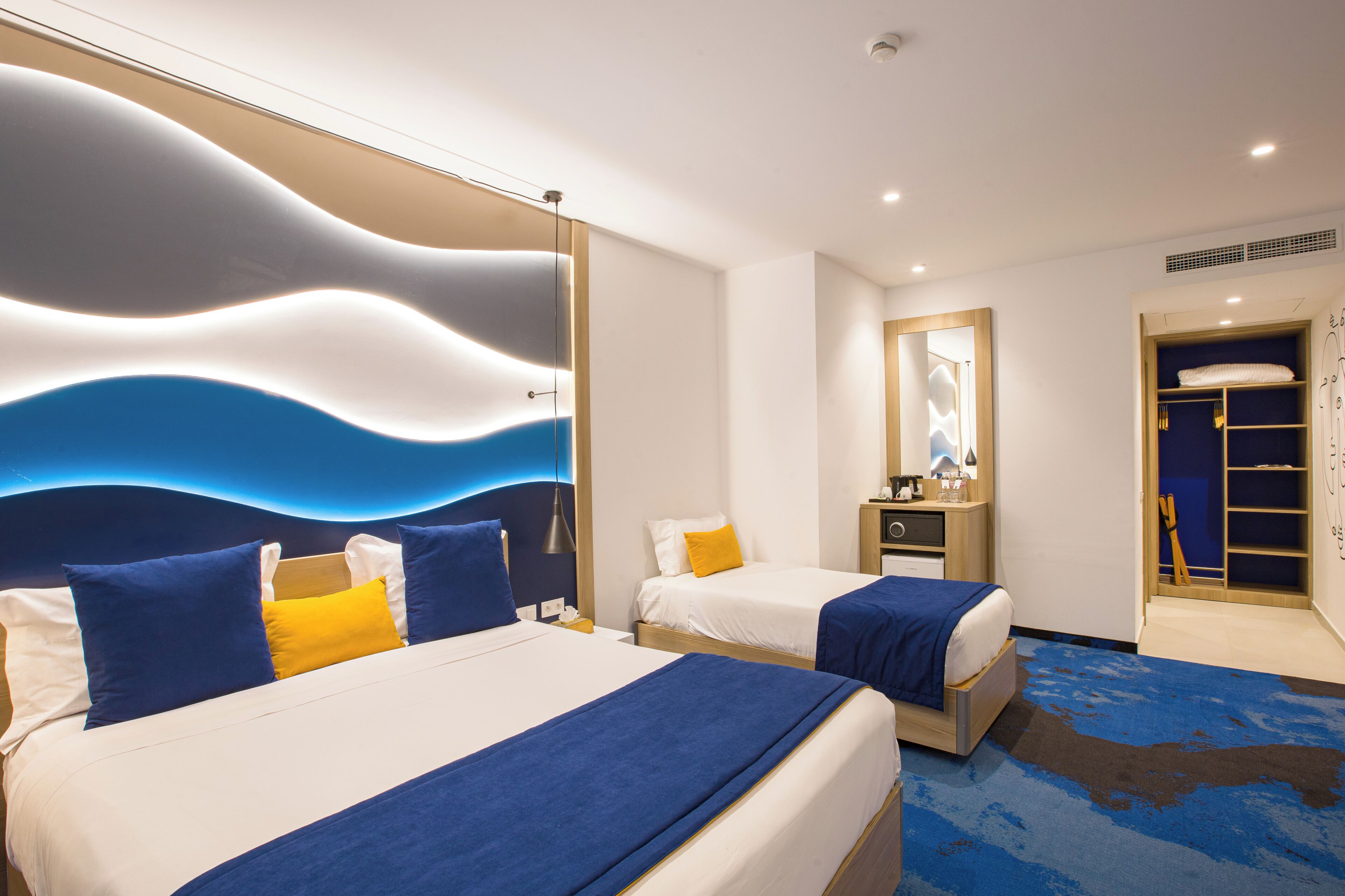 A hotel room with two beds, a large wall mural, a mirror, and a blue carpet.