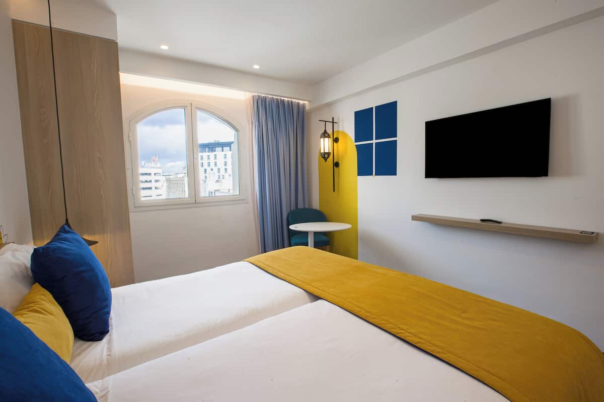 A hotel room with two beds, a television, a window with curtains, and a city view.