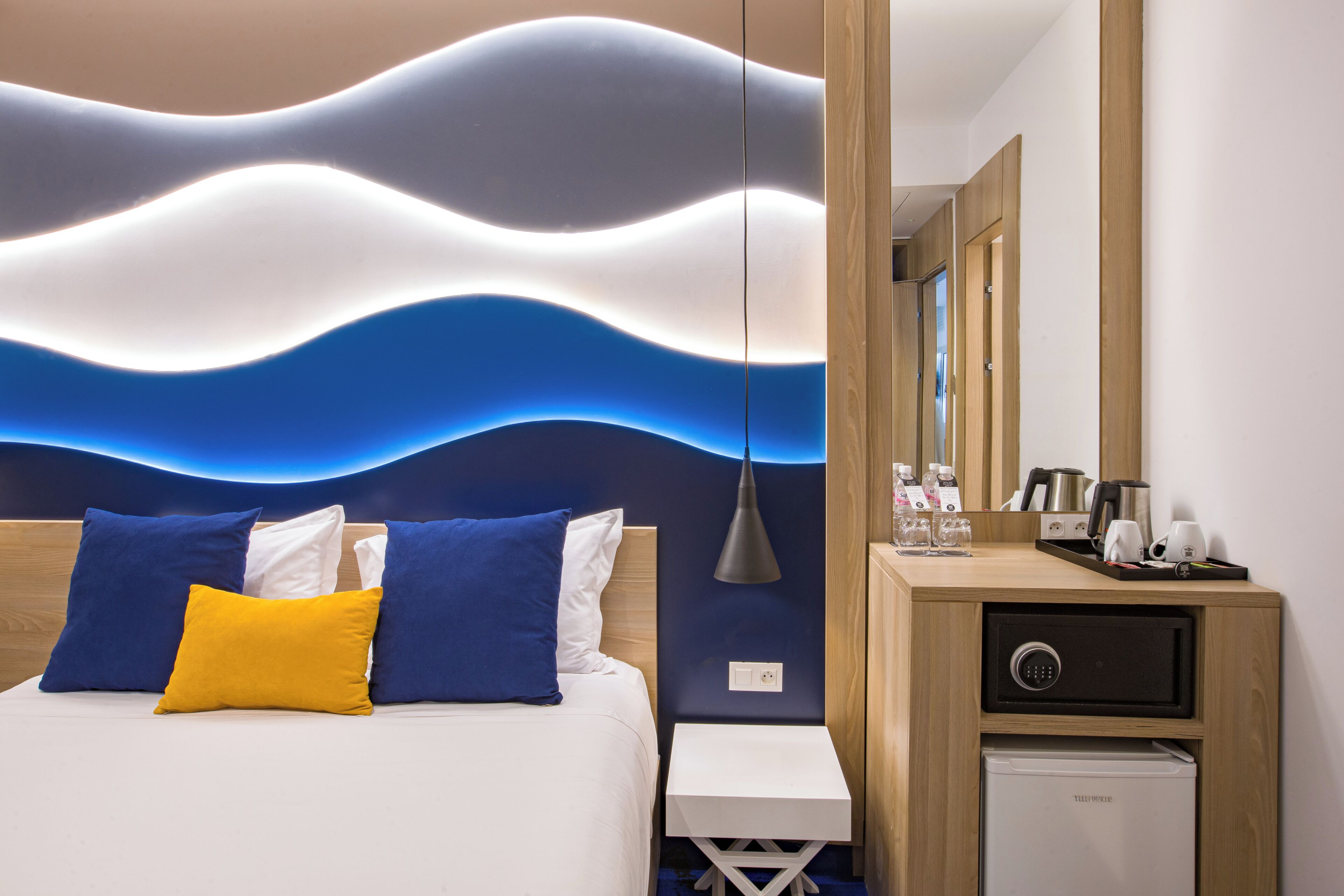 Een hotelkamer met een bed, nachtkastje, klein bureau, kluisje en spiegel.