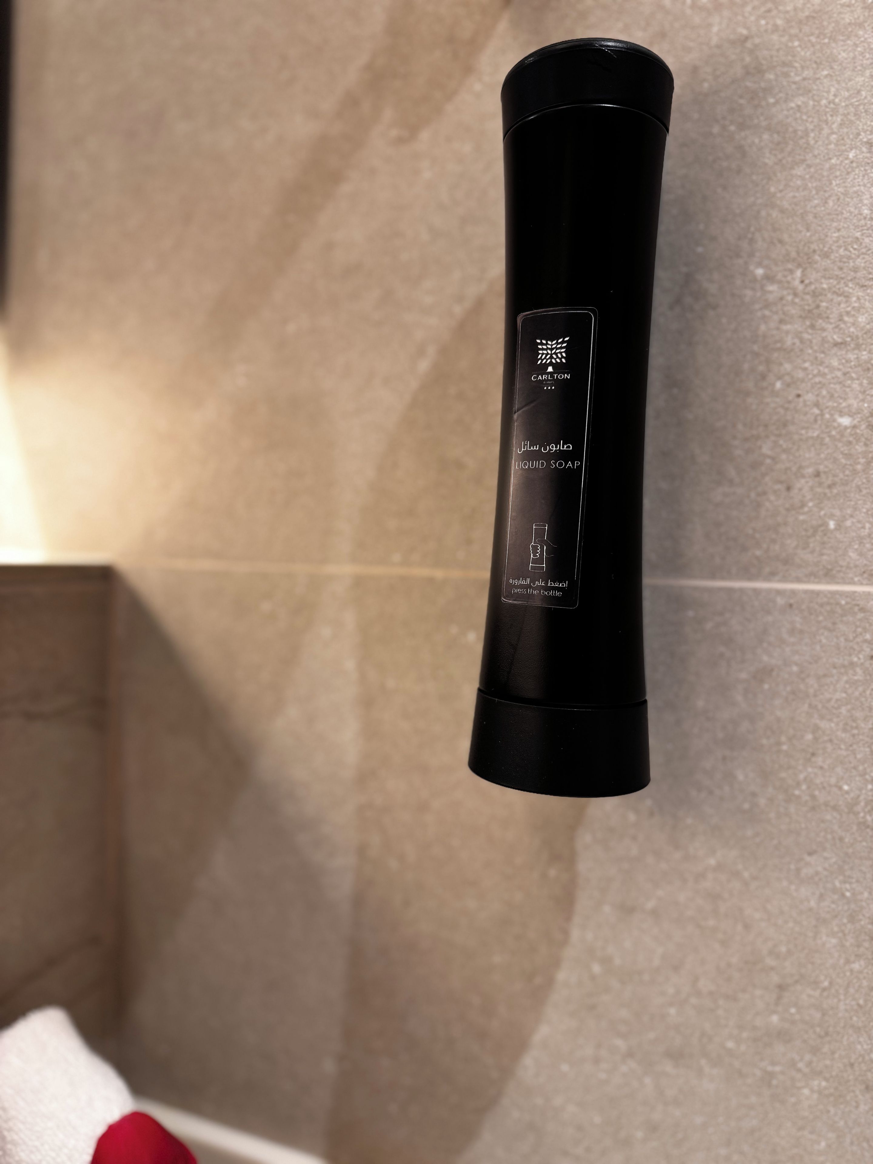 A black cylindrical container labeled 'CARLTON LIQUID SOAP' with instructions to 'press the bottle' on a beige tiled wall.