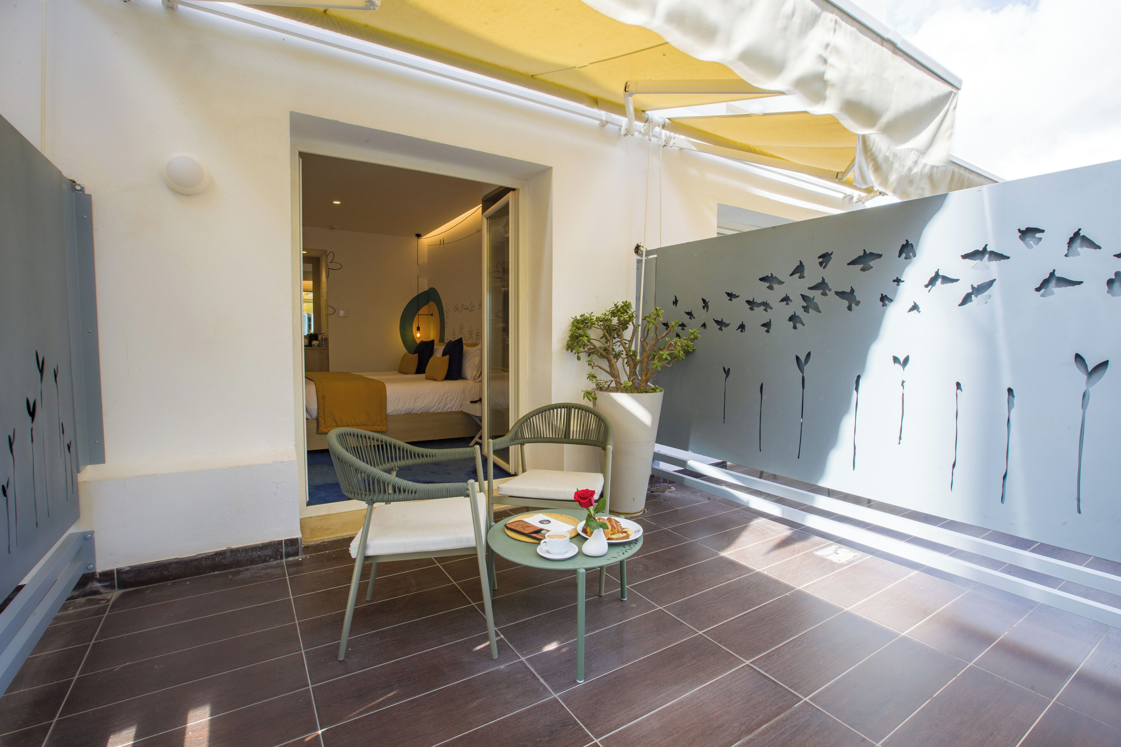 A room with a bed, a chair, a table with a vase and plates, a potted plant, and a wall with bird silhouettes.