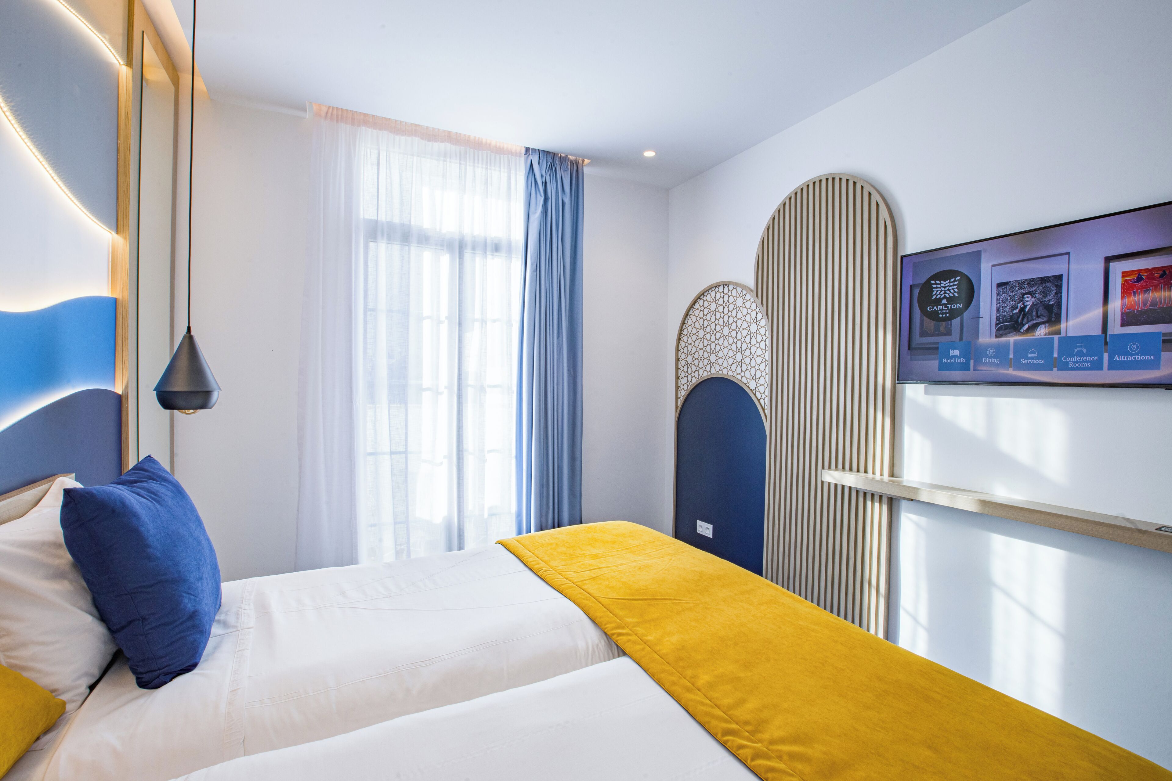 A hotel room with a bed, a window with curtains, a wall-mounted TV, and a decorative panel.