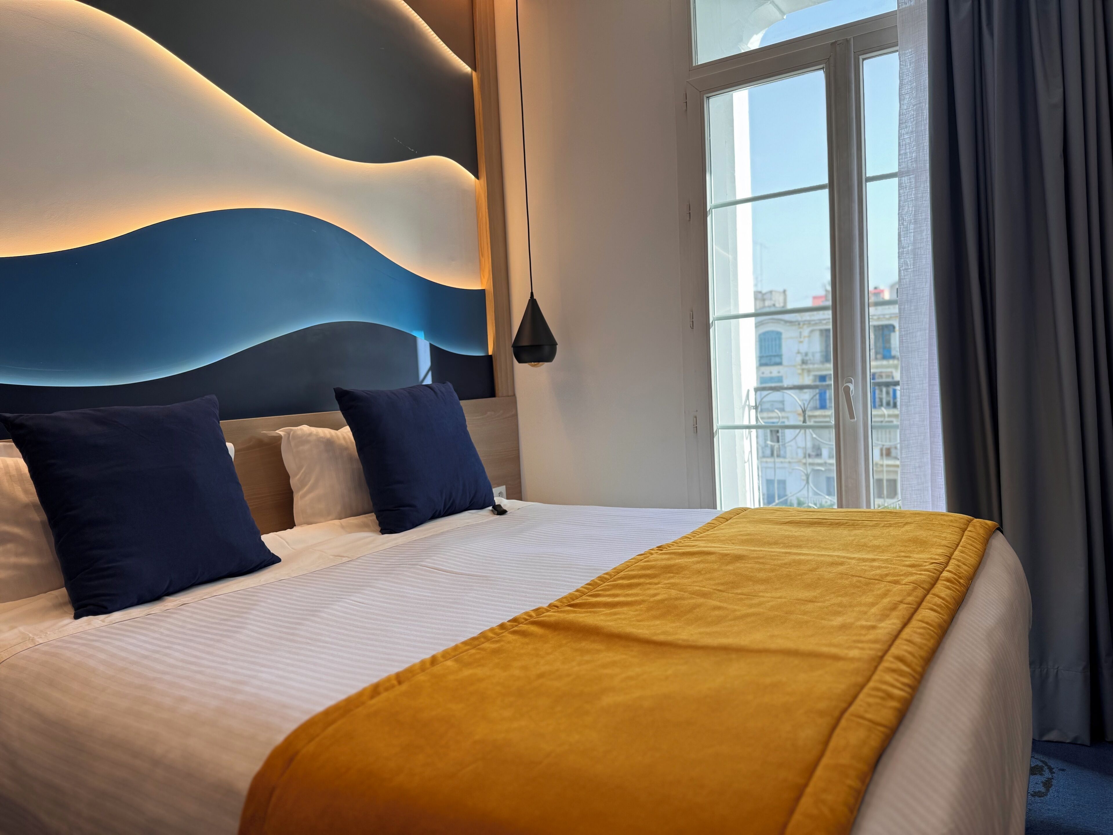 A modern hotel room with a large bed, a view of the city through a large window, and a unique wall design.