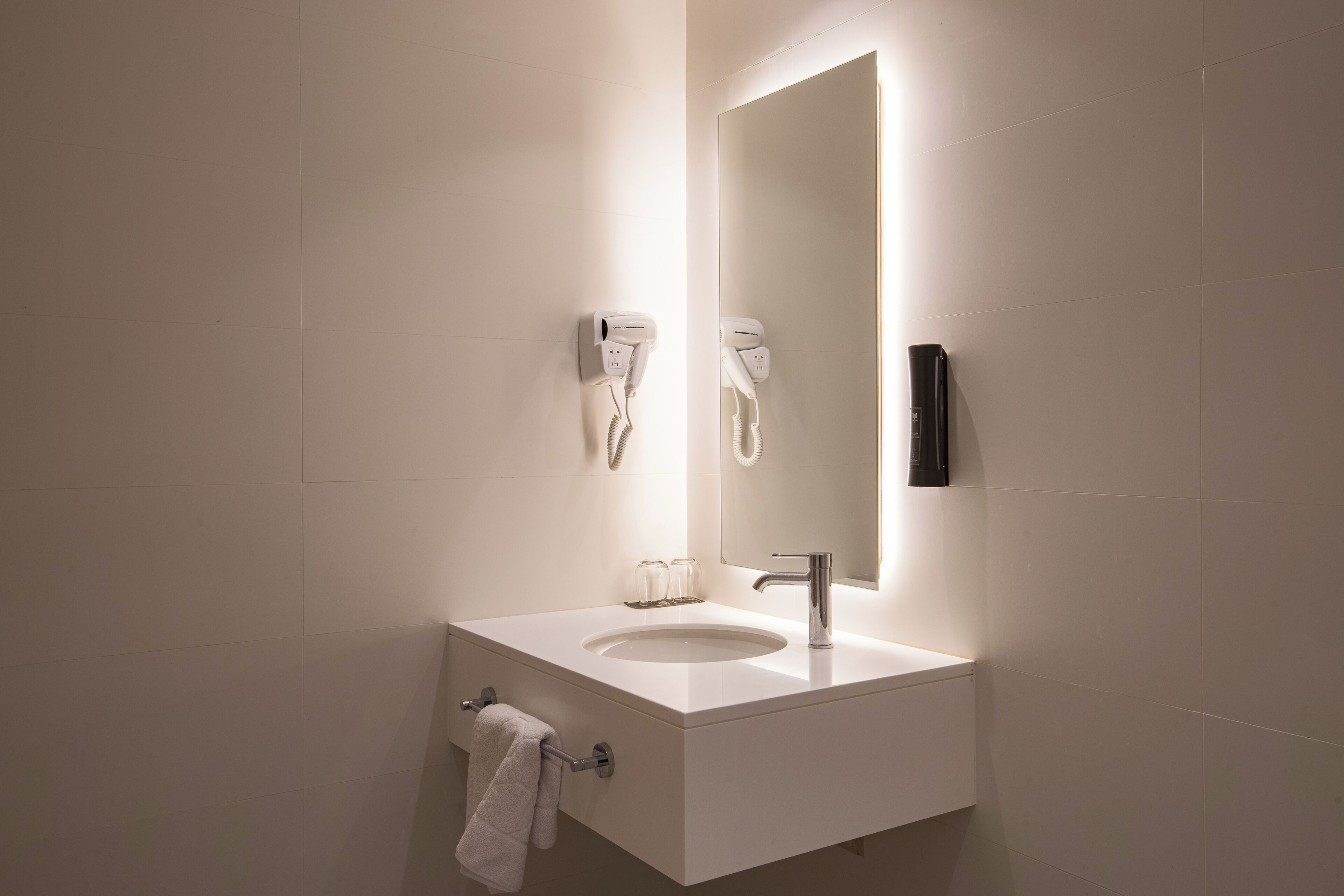 Un baño moderno con un lavabo blanco, un espejo, un perchero para toallas y un secador de pelo fijado a la pared.