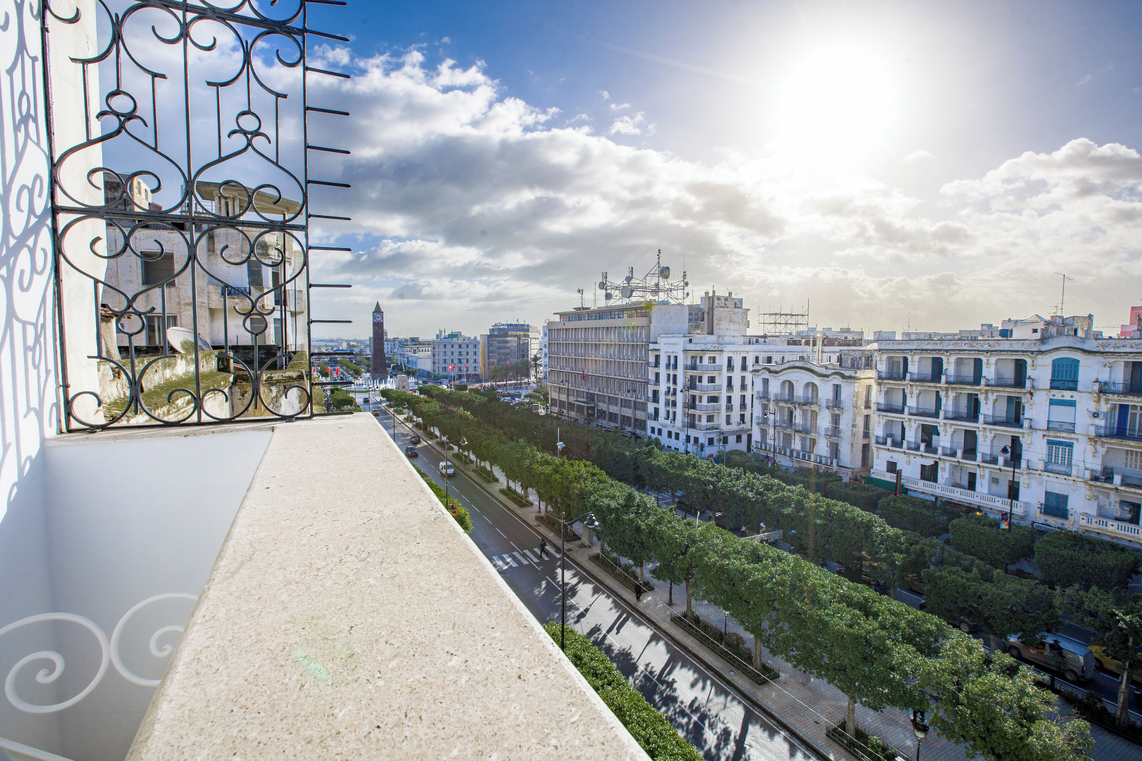 A balcony with a metal railing overlooking a city street with buildings and trees.