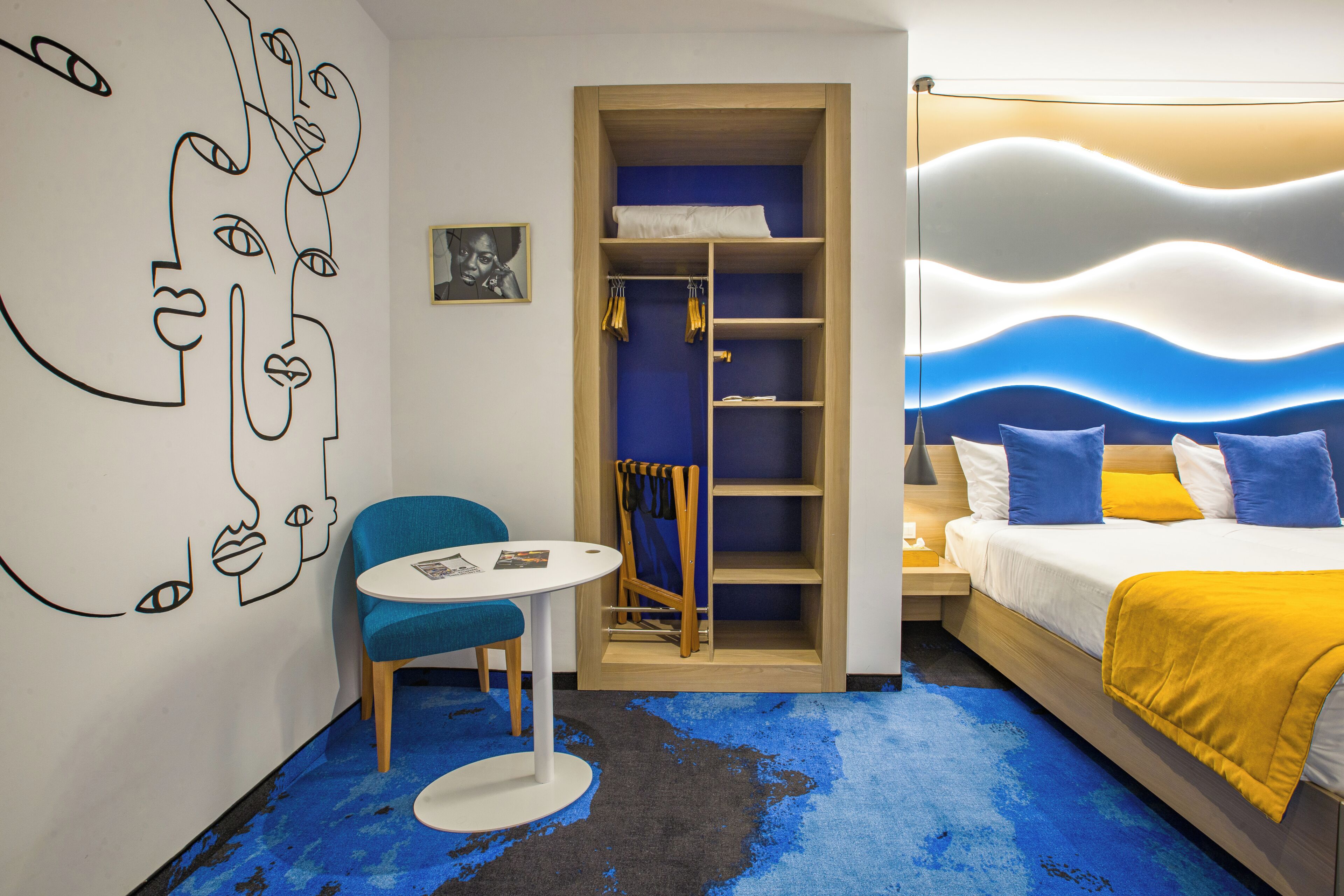 A room with a bed, a chair, a small table, a wardrobe, and a wall with a line drawing.
