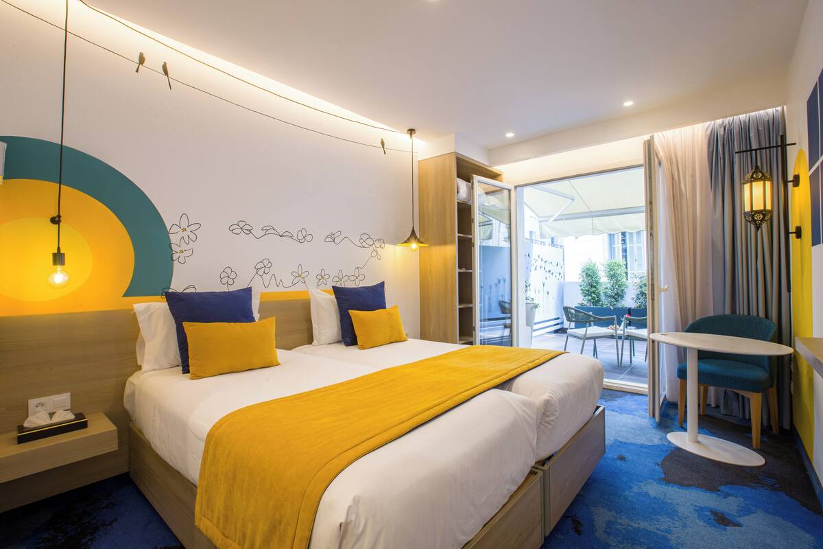 A hotel room with a bed, bedside table, lamp, and a view of the outdoors.