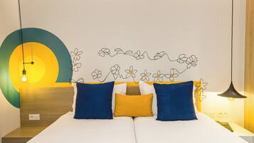 Una cama doble con almohadas azules y amarillas, una pared con diseño circular y motivos florales, y un colgador de luz.