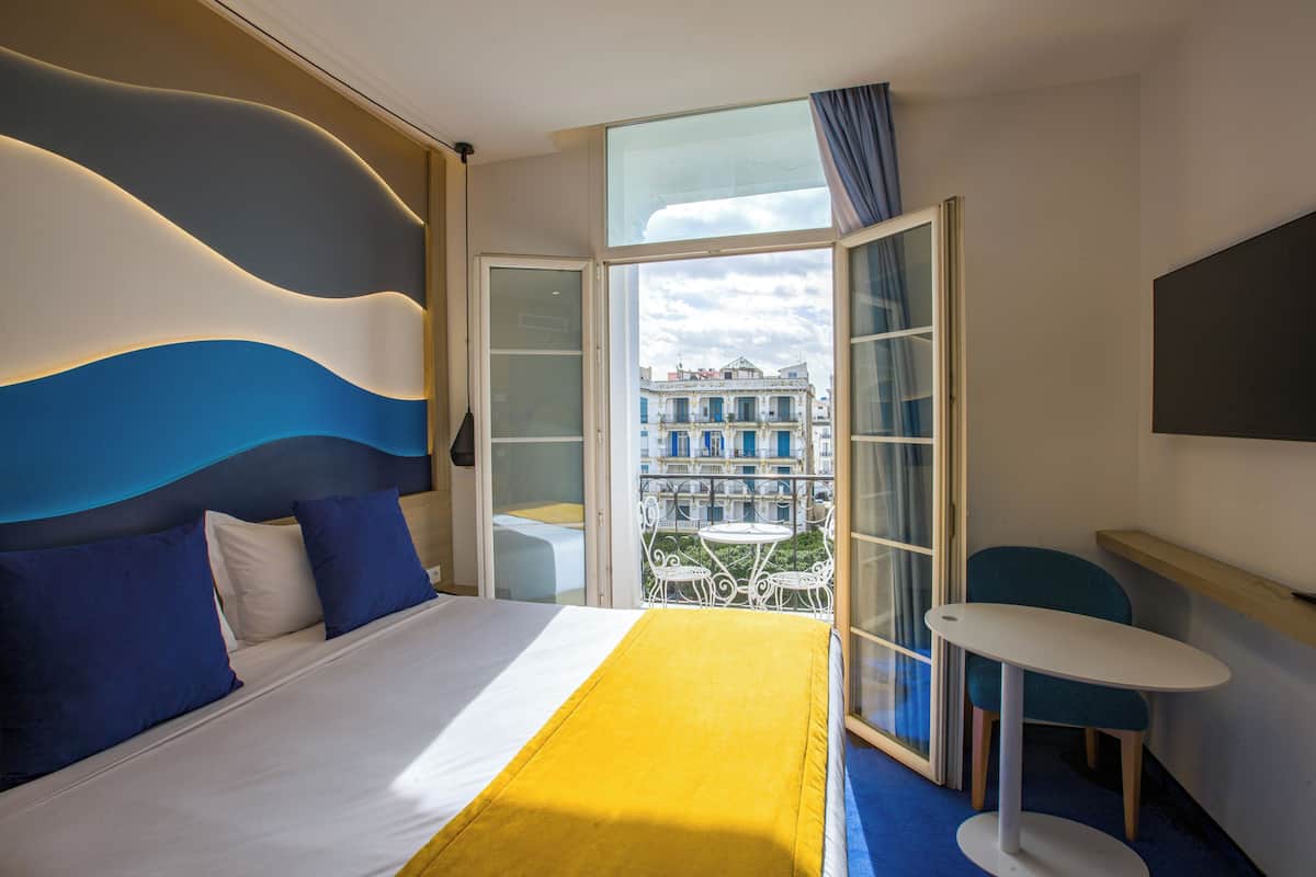 A hotel room with a bed, a balcony with a table and chairs, and a view of buildings.