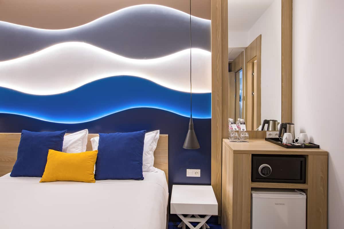 A hotel room with a bed, bedside table, a small desk, a safe, and a mirror.