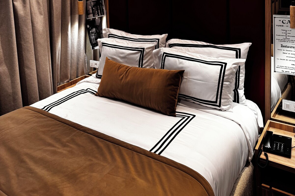 A hotel room with a bed, a nightstand, a telephone, and a sign that reads 'Carlton Tunis Since 1926'.