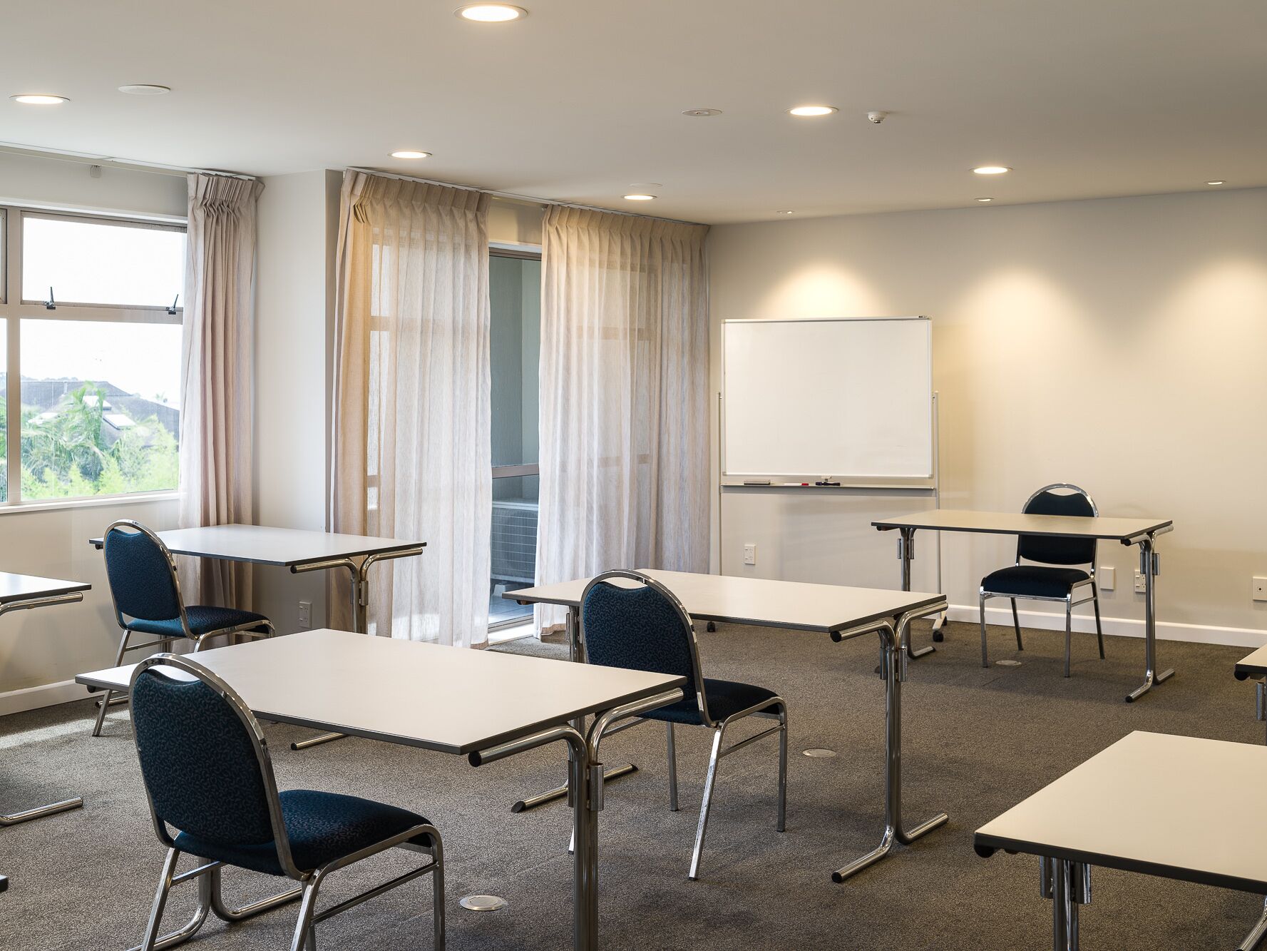 Sala de conferência com mesas, cadeiras, quadro branco e janela com cortinas.