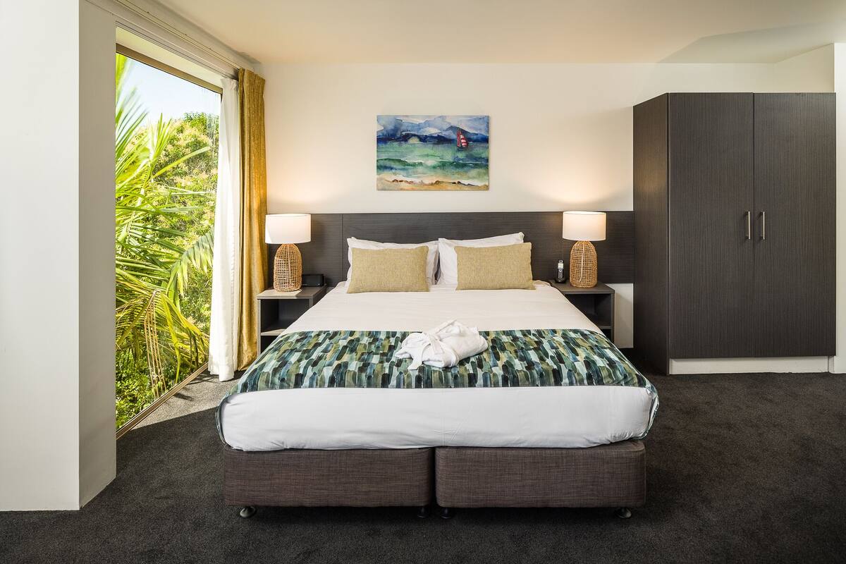 A hotel room with a large bed, two bedside tables, a dark wardrobe, a painting on the wall, and a view of greenery through the window.