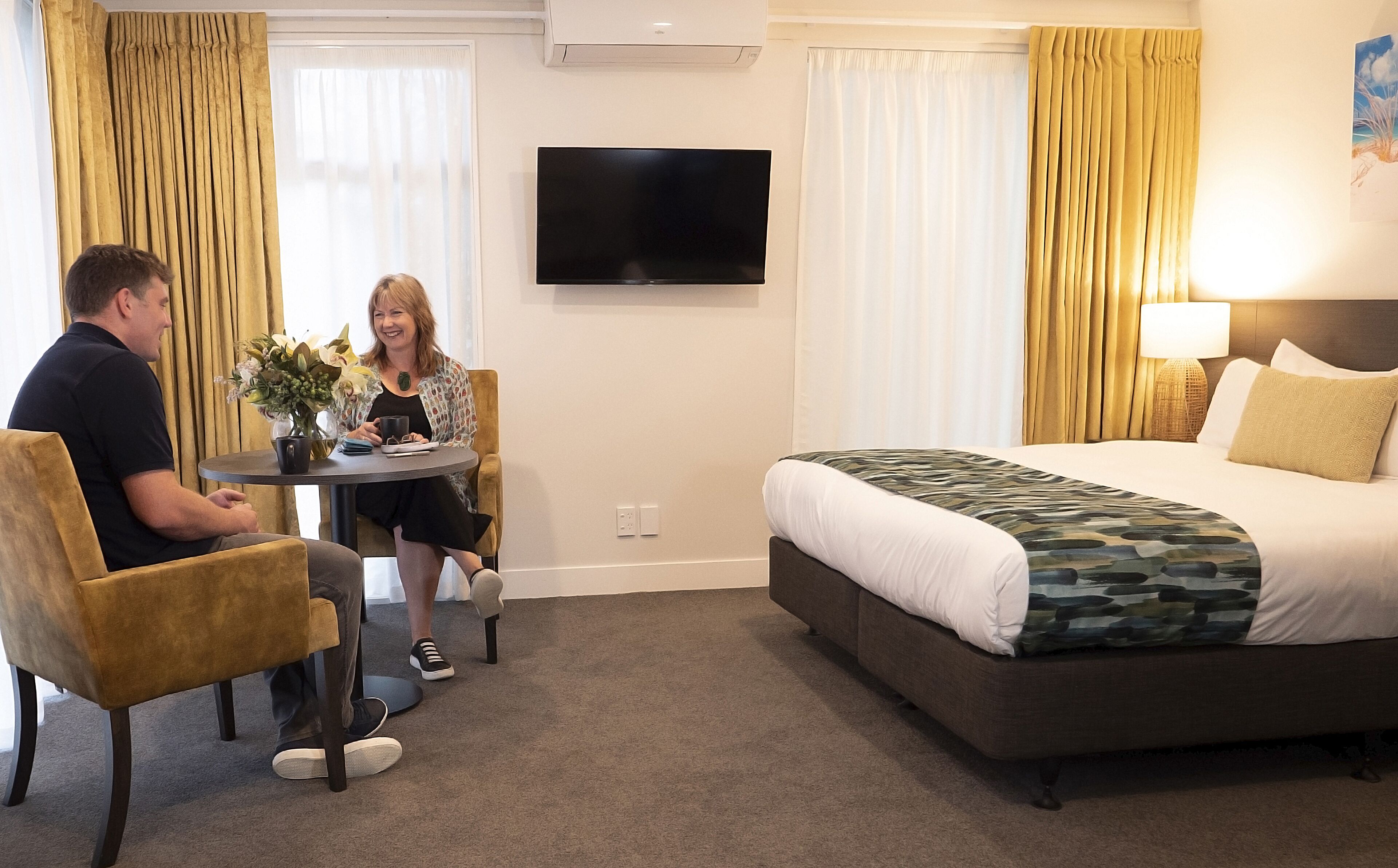Habitación de hotel con cama, televisión, mesa y sillas, y ventana con cortinas.