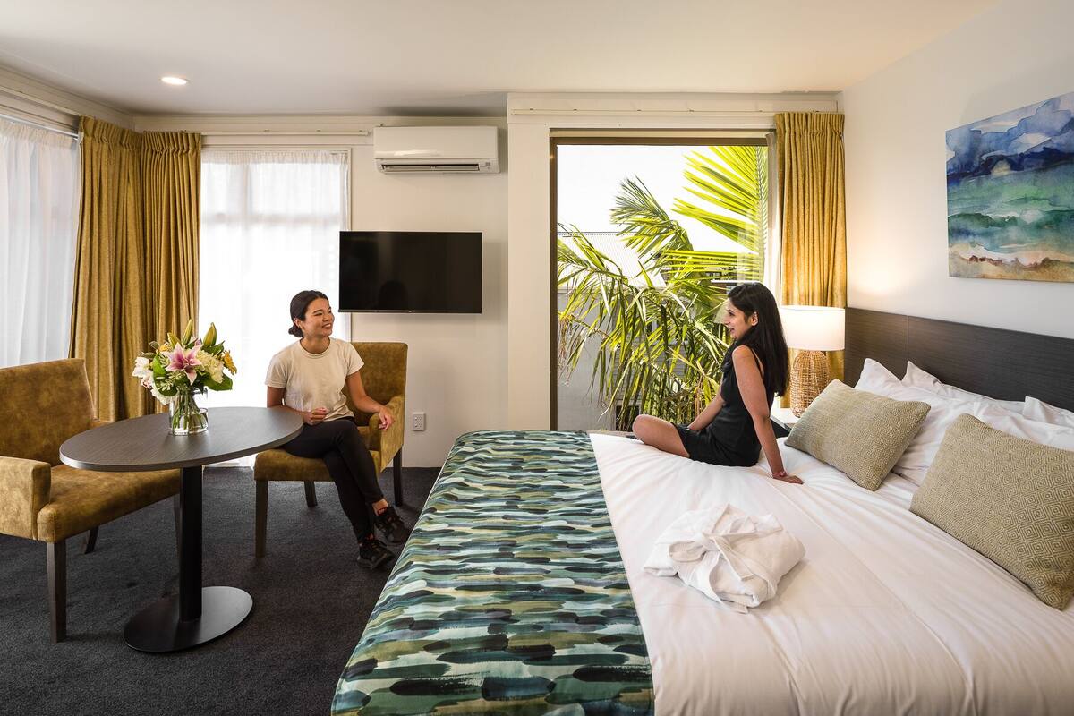 A hotel room with a bed, a sofa, a table with a vase of flowers, and a TV on the wall.