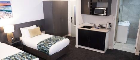 Habitación de hotel con cama, escritorio con lámpara, kitchenette con microondas y zona de ducha.