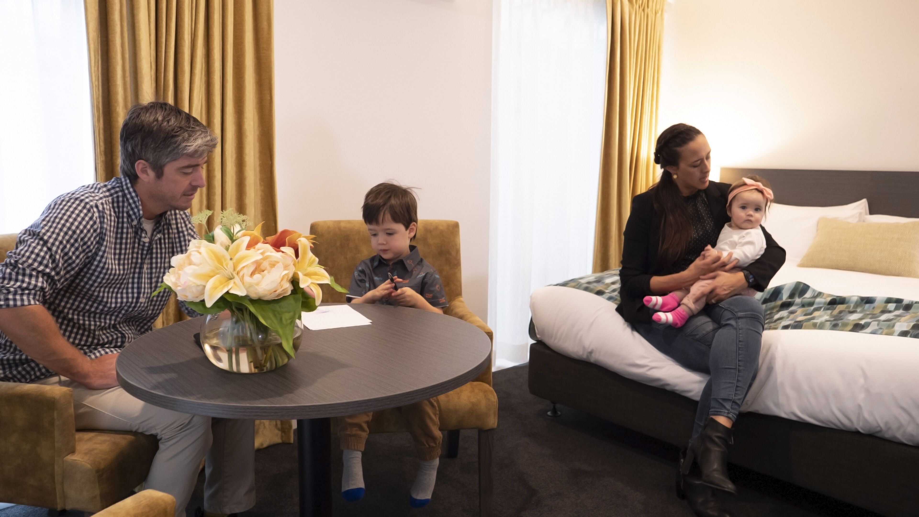 A hotel room with a round table, a bed with a patterned bedspread, and a vase of flowers on the table.