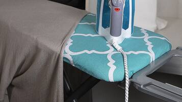 A neatly organized room with a bed, an ironing board with an iron, a robe hanging on a rack, and a control panel on the wall.