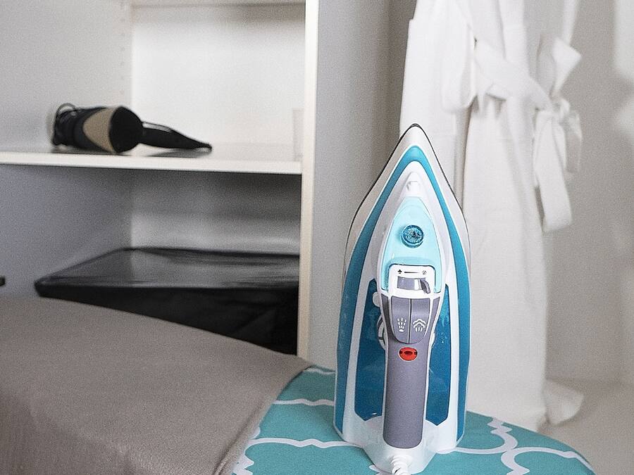 A neatly organized room with a bed, an ironing board with an iron, a robe hanging on a rack, and a control panel on the wall.