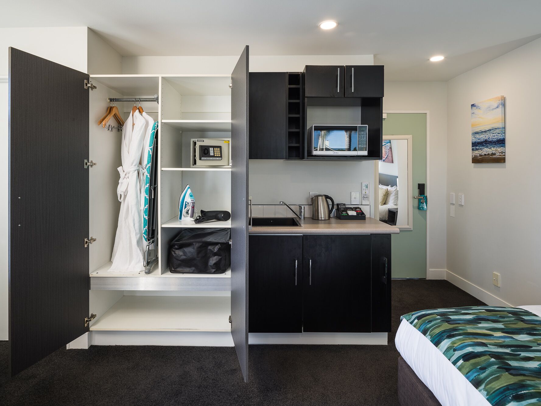 A compact hotel room with a kitchenette, a bed, a wardrobe, and a painting on the wall.