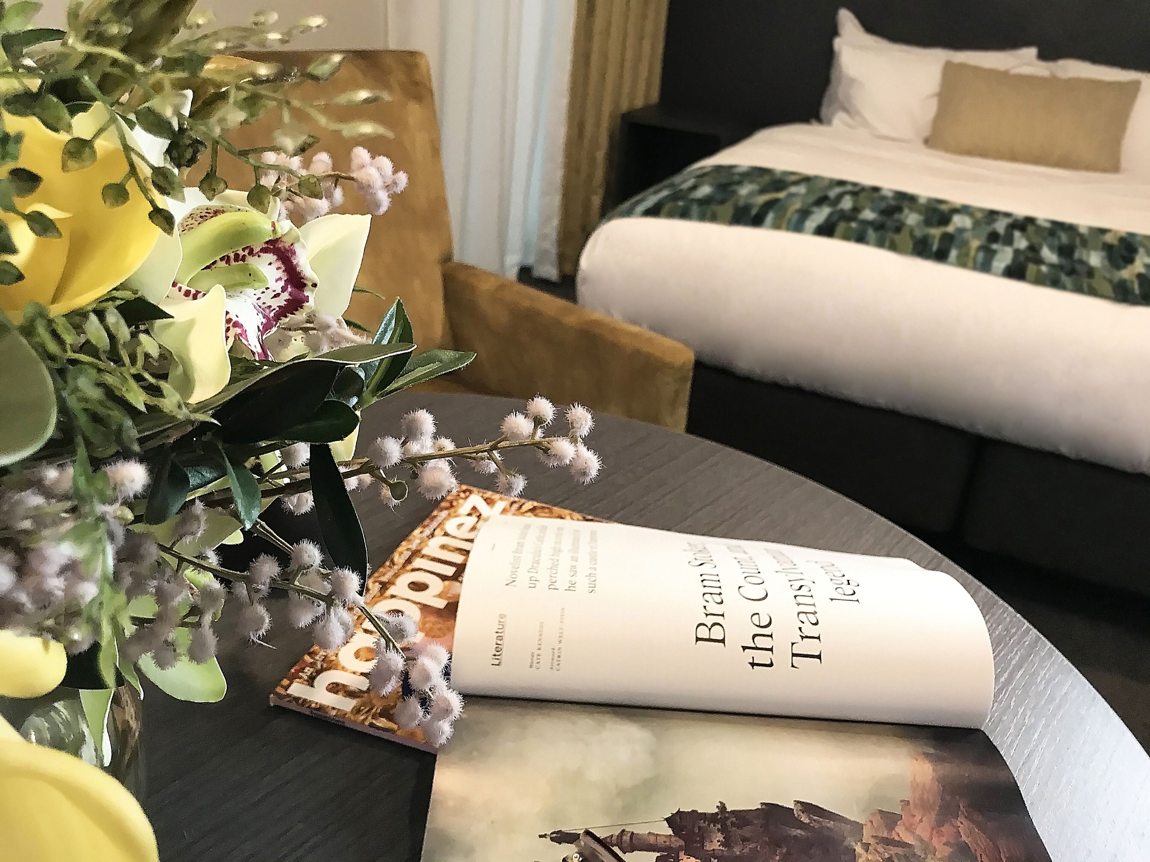 A hotel room with a bed, a bedside table with a book and a vase of flowers.