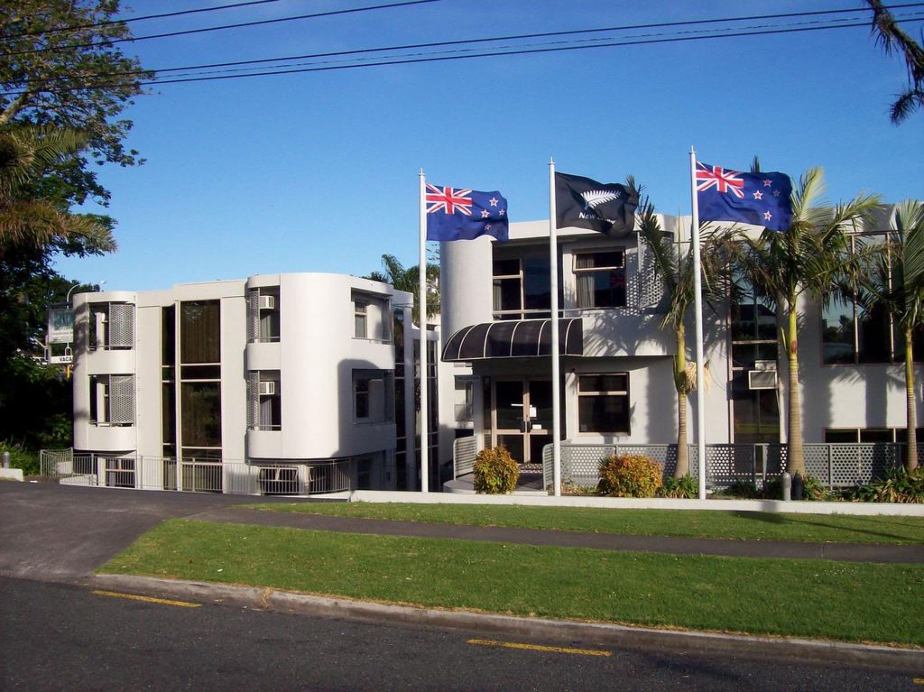 Un edificio moderno con varias banderas, incluyendo las de Australia y Nueva Zelanda, y un césped bien cuidado.