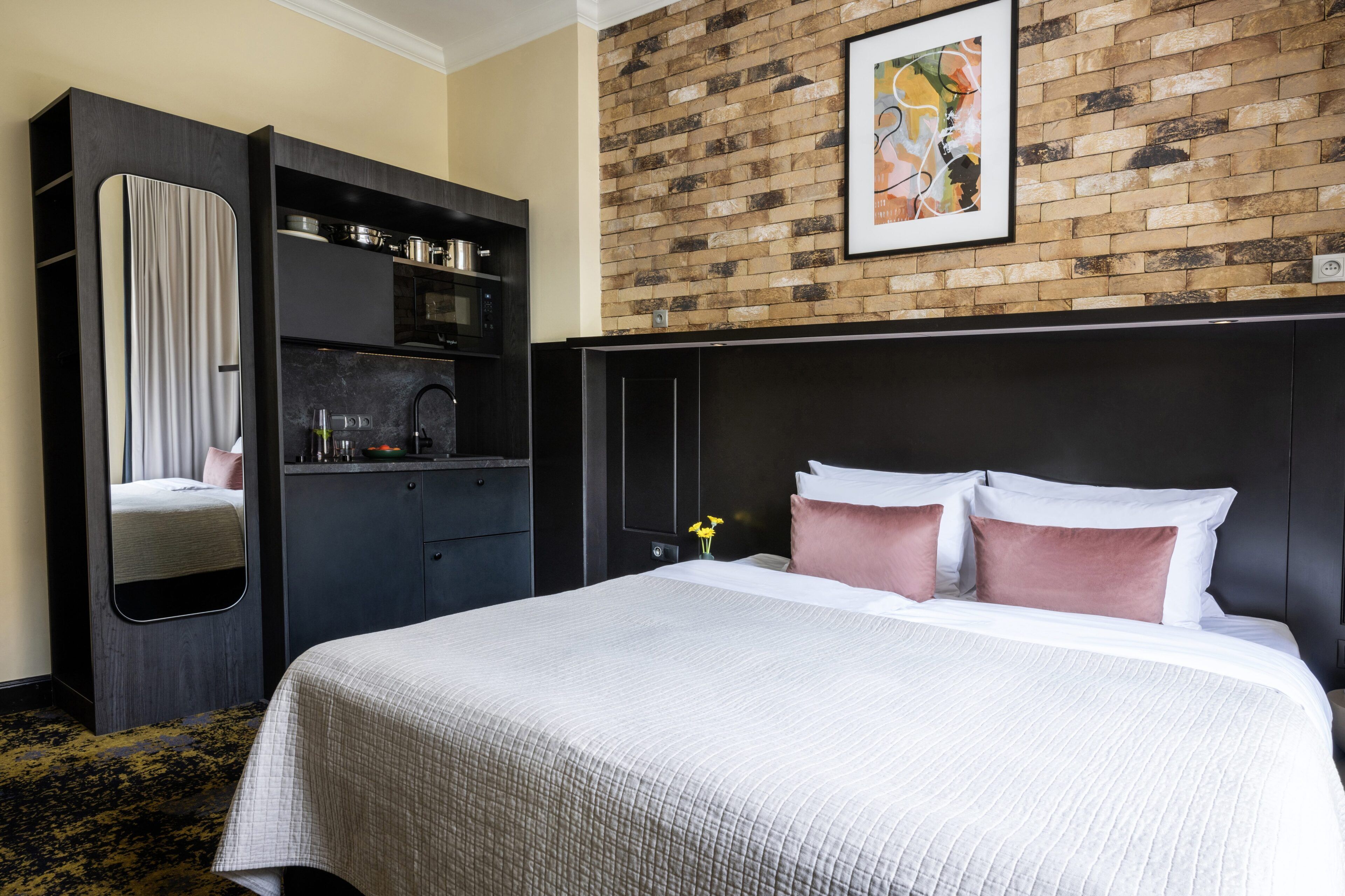 A hotel room with a bed, a wardrobe, a mirror, a brick wall, and a painting.