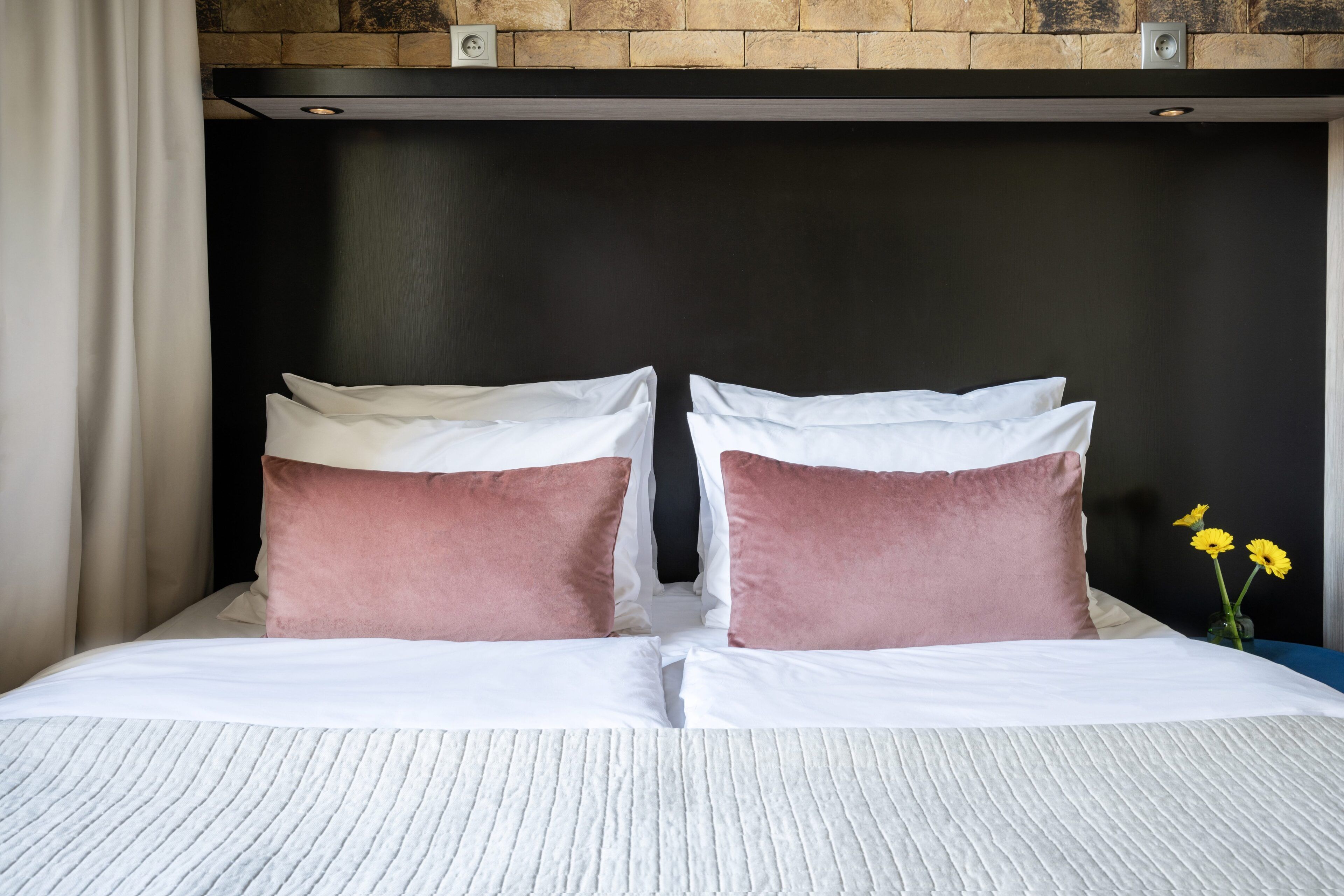 A bed with white linens and two pillows, one pink and one white, in a room with a brick wall and a black headboard.