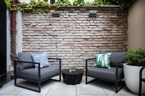 A patio area with two grey sofas, one with a blue pillow and the other with a green pillow, a small round table with a glass and a drink, and a large potted plant. - Numa Prague Root (Prague)