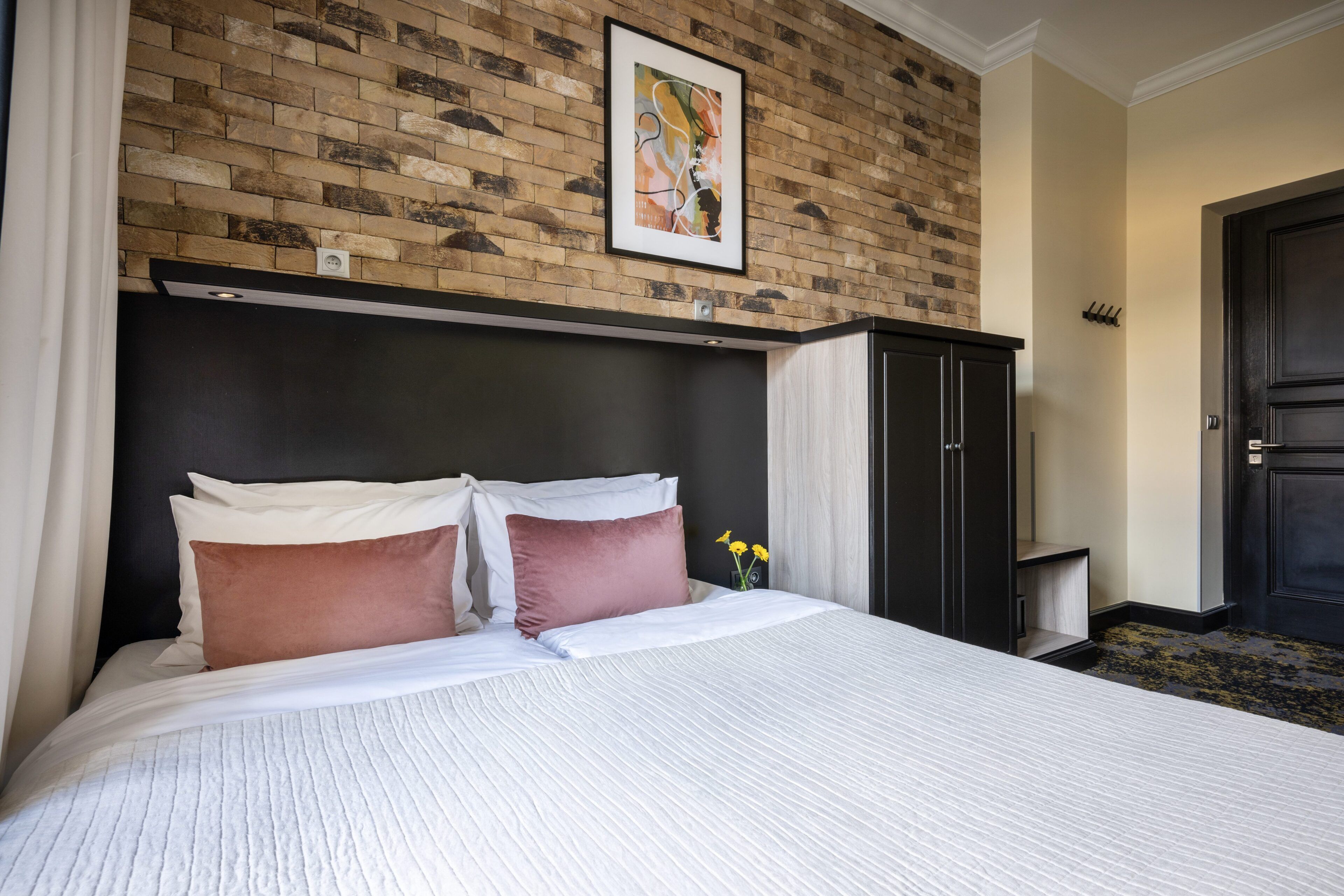 A hotel room with a brick-patterned wall, a bed with white and brown bedding, a black wardrobe, and a door with a coat rack.