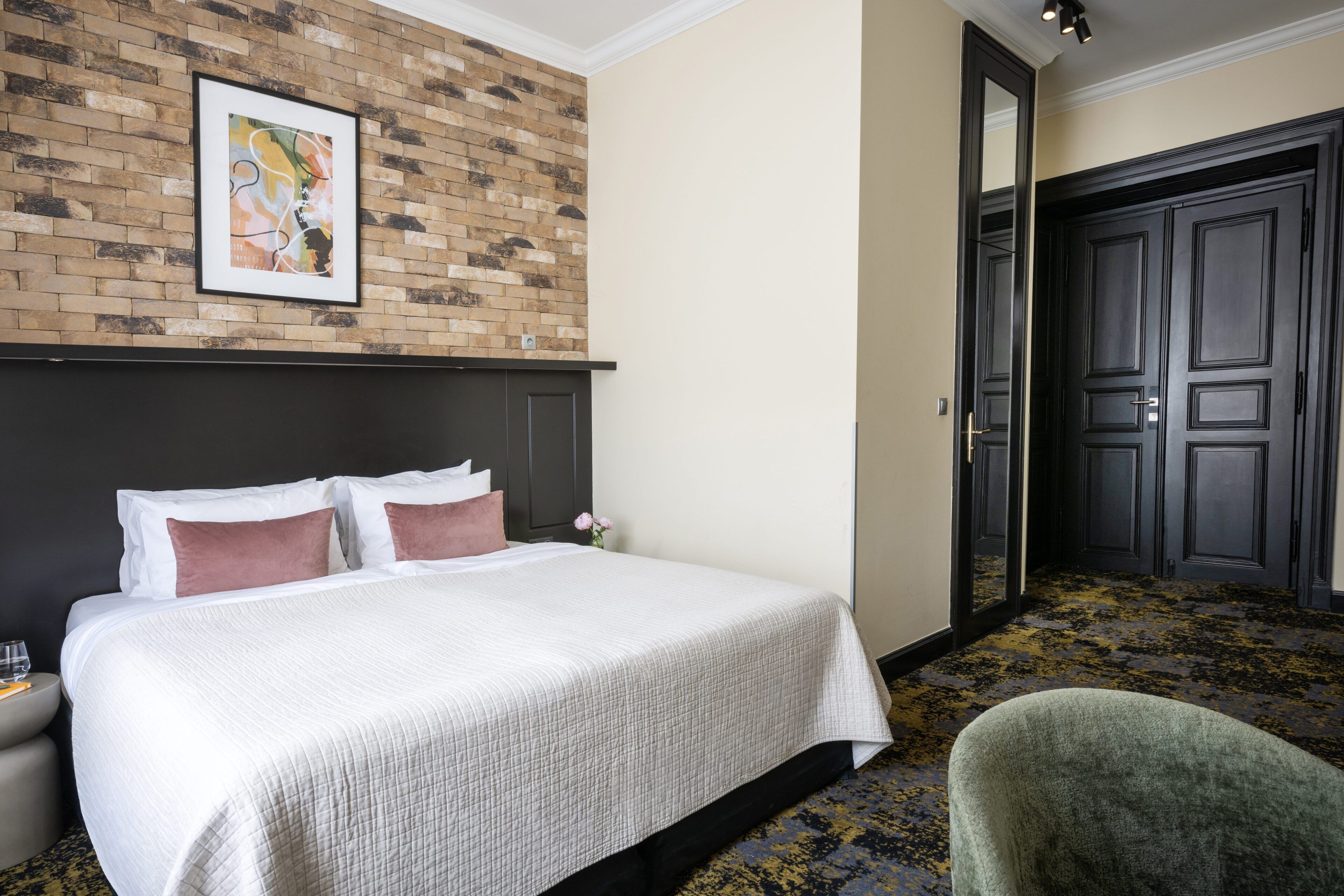 A hotel room with a bed, a nightstand, a chair, a brick wall, and a door.