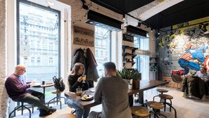 Una cafetería con mesas y sillas, ventanas amplias y un mural en la pared.