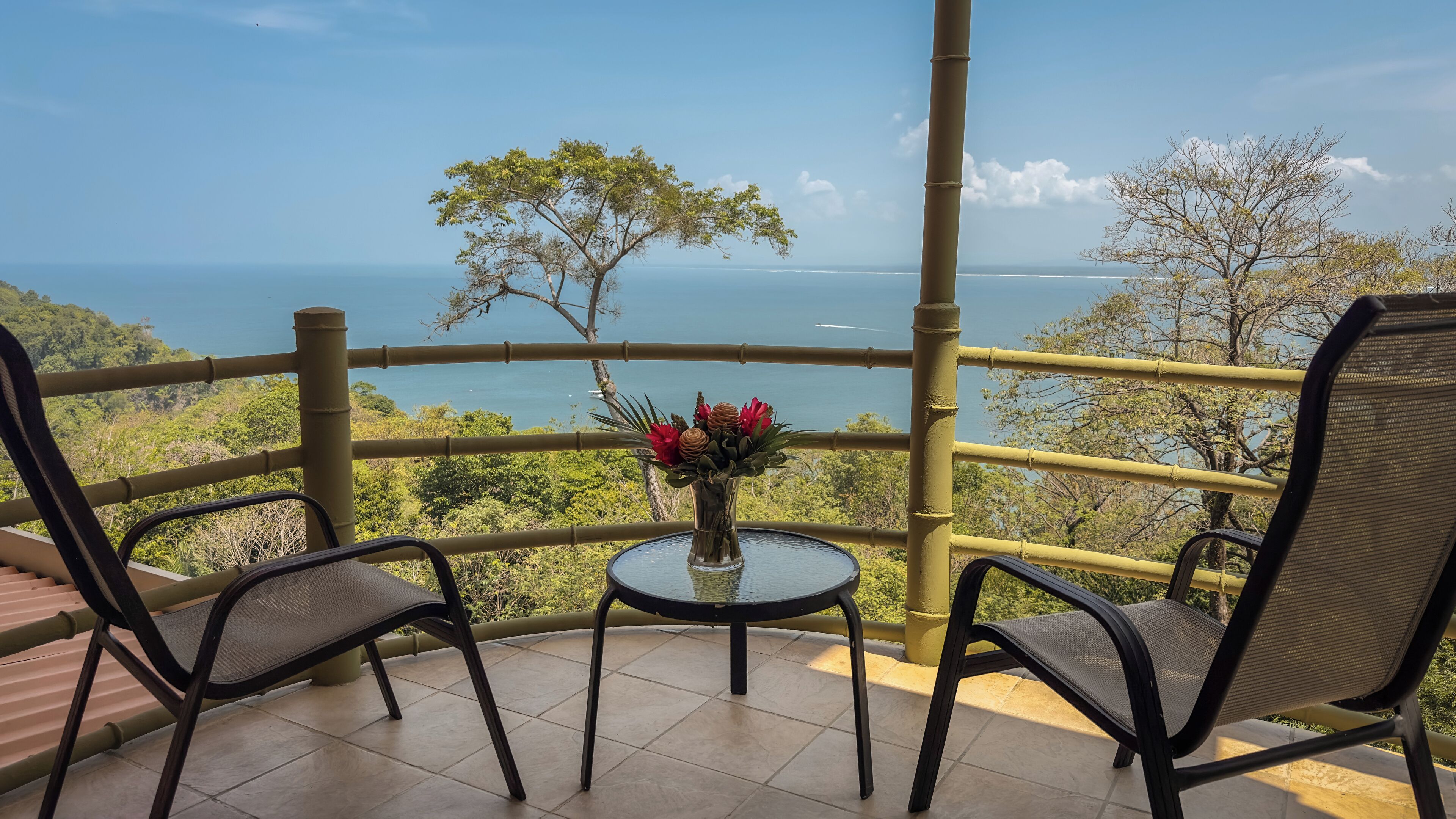 Un balcón con dos sillas y una mesa, con vistas a un paisaje marino y abundante vegetación.